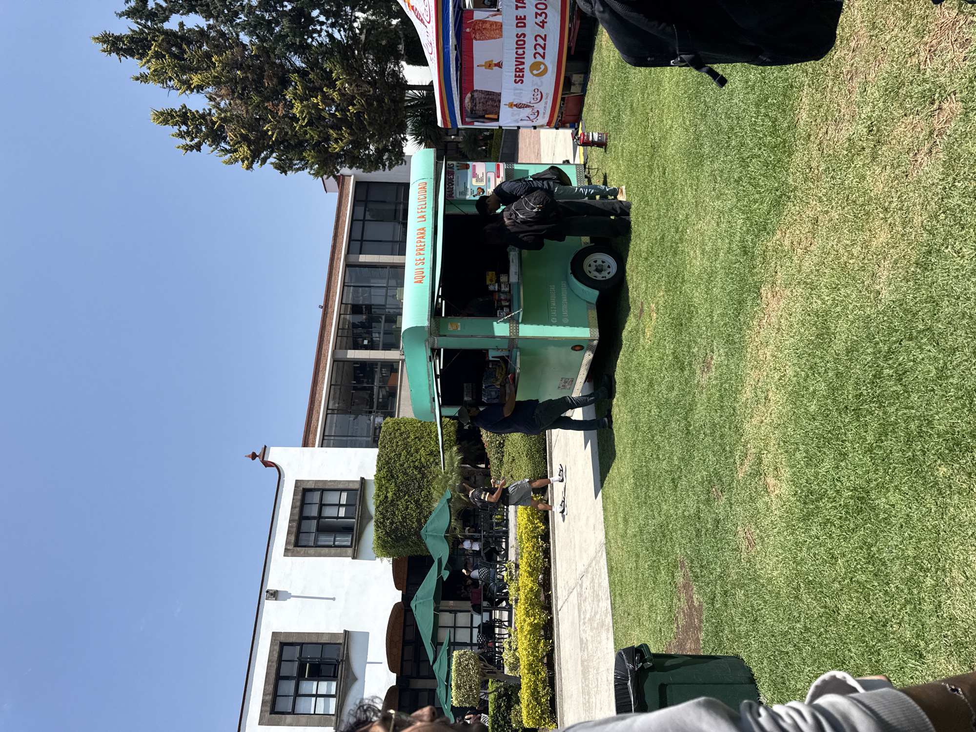 An outdoor scene captured on a bright, sunny day shows a vibrant gathering with food vendors. The primary focus is a light blue/teal food truck with a serving window, where several people are seen standing, possibly ordering or waiting for food. The side of the food truck bears the Spanish text "AQUI SE PREPARA LA FELICIDAD" (Here happiness is prepared), along with "LAS 3 MARQUESAS" and the social media handle "@LASTRESMARQUESAS".
Adjacent to the food truck is another vendor setup, featuring a white and red tent or stall. Visible text on this stall includes "SERVICIOS DE TA", "LO MEJOR DEL TACO" (The best taco), "222 430-", and a logo that appears to say "K-aco".
The food vendors are positioned on a paved pathway next to a well-maintained lawn of green grass. In the background, there's a white building with dark-framed windows and a part of a building with a red roof. Several green patio umbrellas and outdoor tables suggest a seating area for patrons. Sparse trees are visible against the clear blue sky, casting distinct shadows, indicating a high sun position. People are casually strolling or standing in the area, contributing to a lively atmosphere. A dark green trash can is visible in the foreground on the right.