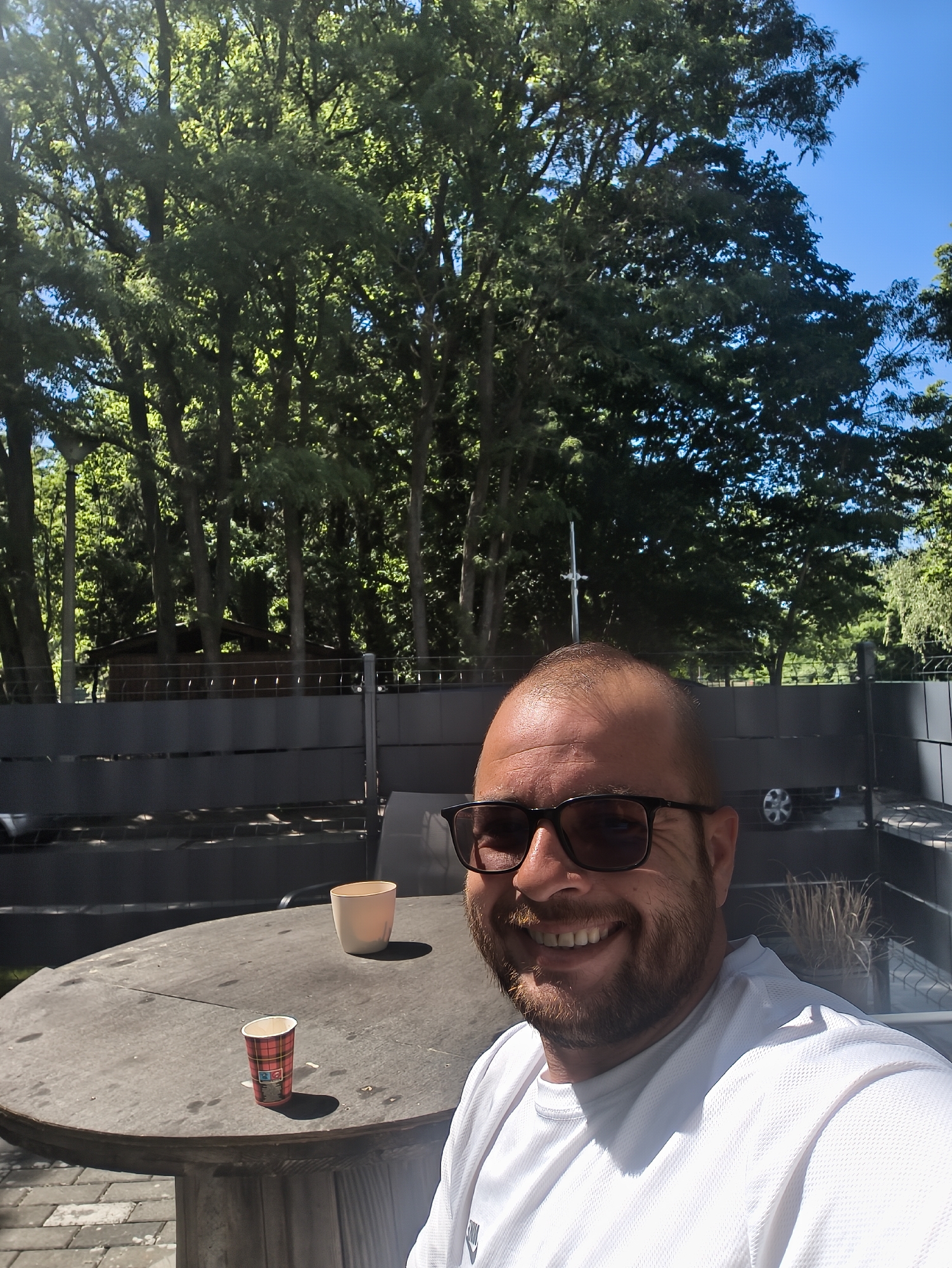 The image is a selfie of a smiling man with a beard and sunglasses. He is wearing a white shirt. The background shows a gray table with two cups on it, a gray fence, trees, and a blue sky. There's a glimpse of a car parked behind the fence. The location appears to be an outdoor patio or backyard. The city and country are not identifiable from the image.