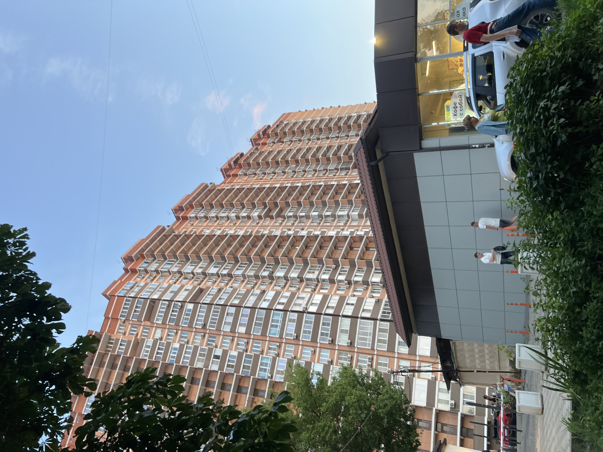 In Krasnodar, Russia, a tall, multi-story brick apartment building with numerous balconies dominates the skyline. In the foreground, a low-rise commercial building with a gray facade and a dark roof is situated. Several individuals are visible: two men stand near the commercial building, one holding a black backpack; another man walks past them carrying a shopping bag. A woman wearing a light blue jacket and a yellow top is near a silver SUV, partially obscured by the vehicle. A man in a red shirt and blue jeans is seen walking away from the SUV. A red car is parked in the background. The scene appears to be a street or public area with orange traffic cones and planters filled with green foliage. The sky is clear with a few wispy clouds.