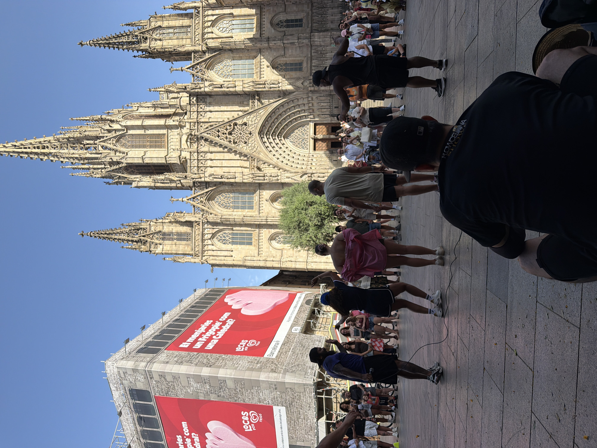 This media file shows a street scene in Barcelona, Spain, in front of a large Gothic cathedral. A diverse crowd of people, many appearing to be tourists or locals enjoying a sunny day, are gathered on a stone-paved plaza. Some individuals are watching or participating in an activity, possibly a performance or demonstration, as suggested by the presence of a microphone and a cord snaking across the ground.
Dominating the background is the majestic facade of the cathedral, with its intricate spires and detailed stonework. Adjacent to the cathedral is a building covered in large red banners featuring images of feet and text in Catalan, including phrases like "El menjaries un Frigopie com una Catedral?" (Would you eat a Frigopie like a Cathedral?) and logos for "LOCOS POR FRIGO" and "Barcelona." This suggests a promotional event or advertisement campaign is taking place.
The time of day appears to be midday or early afternoon, given the bright sunlight casting distinct shadows. The weather seems pleasant and warm, contributing to the outdoor gathering. The overall atmosphere seems lively and engaged, with people of various ages and backgrounds interacting and observing. The presence of a microphone implies a potential performance or a spoken presentation to the crowd.