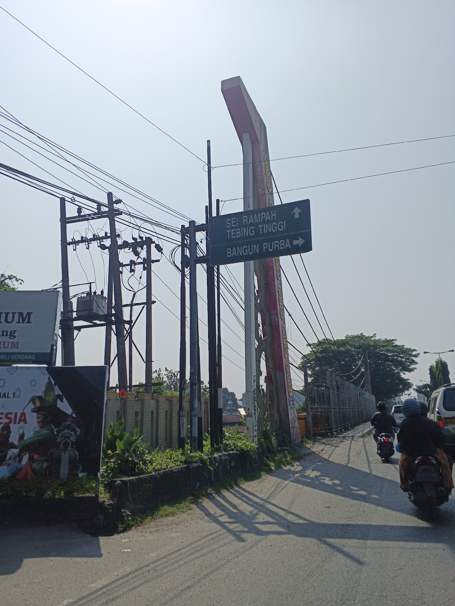 A sunny, clear day in Tg Morawa A, Indonesia, is depicted with a road and its surroundings. The scene is dominated by a road that curves to the right, casting long shadows from overhead power lines and a large advertising billboard on the left.
A prominent, multi-poled electrical transformer sits on the left side of the road, surrounded by a dense network of power lines. To the left of the road, a large, vertical billboard features text and images, including what appears to be a cultural depiction with figures in traditional attire.
A signpost stands near the center of the image, displaying directional information. It indicates "SEI RAMPAH TEBING TINGGI" with an upward arrow and "BANGUN PURBA" with a rightward arrow. This signpost is attached to a decorative pole with red and gold accents.
Several people on motorcycles are traveling along the road. One rider is visible in the foreground, while two others are further down the road. A white van is also present on the right side of the road, heading in the same direction as the motorcycles.
The background features a dense line of trees and a metal fence, with a street lamp visible in the distance. The overall environment suggests an urban or semi-urban area with active infrastructure and traffic.