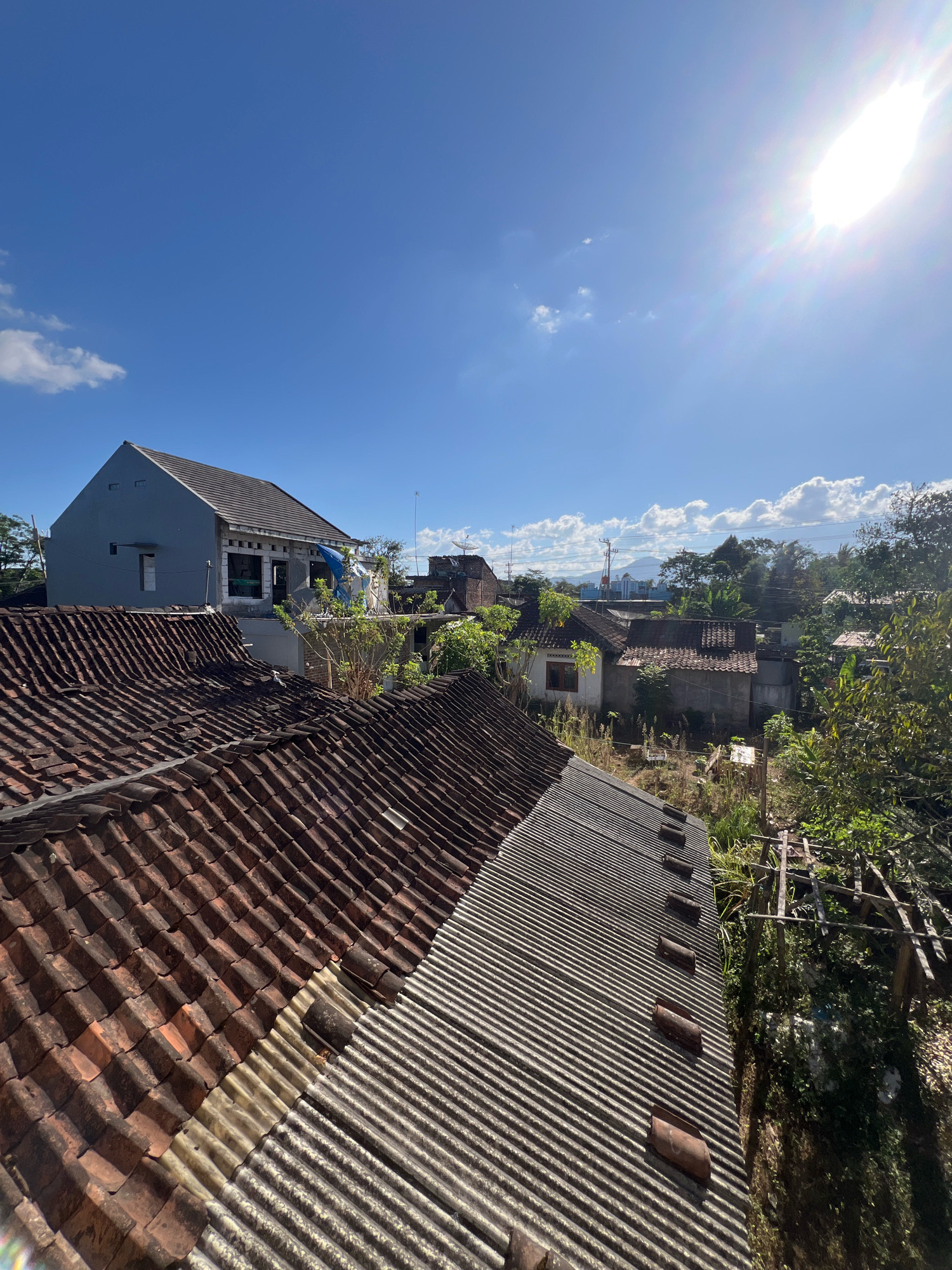 The image shows a rural or suburban residential area under a bright blue sky with some clouds. In the foreground, there are two roofs, one made of reddish-brown terracotta tiles and another of corrugated metal. The terracotta roof appears older and somewhat weathered. The corrugated metal roof shows a row of brown, curved roof tiles placed on top of the metal sheeting. Behind these roofs, a two-story house with light blue siding is visible. Further in the background, more houses, trees, and vegetation are scattered across the landscape. A distant mountain range can be seen on the horizon. The sun is shining brightly in the upper right corner of the frame, casting lens flare. The scene depicts a sunny day in a densely packed residential area with varying architectural styles and roof materials. The overall impression is of a warm, sunny day in a tropical or sub-tropical region.
