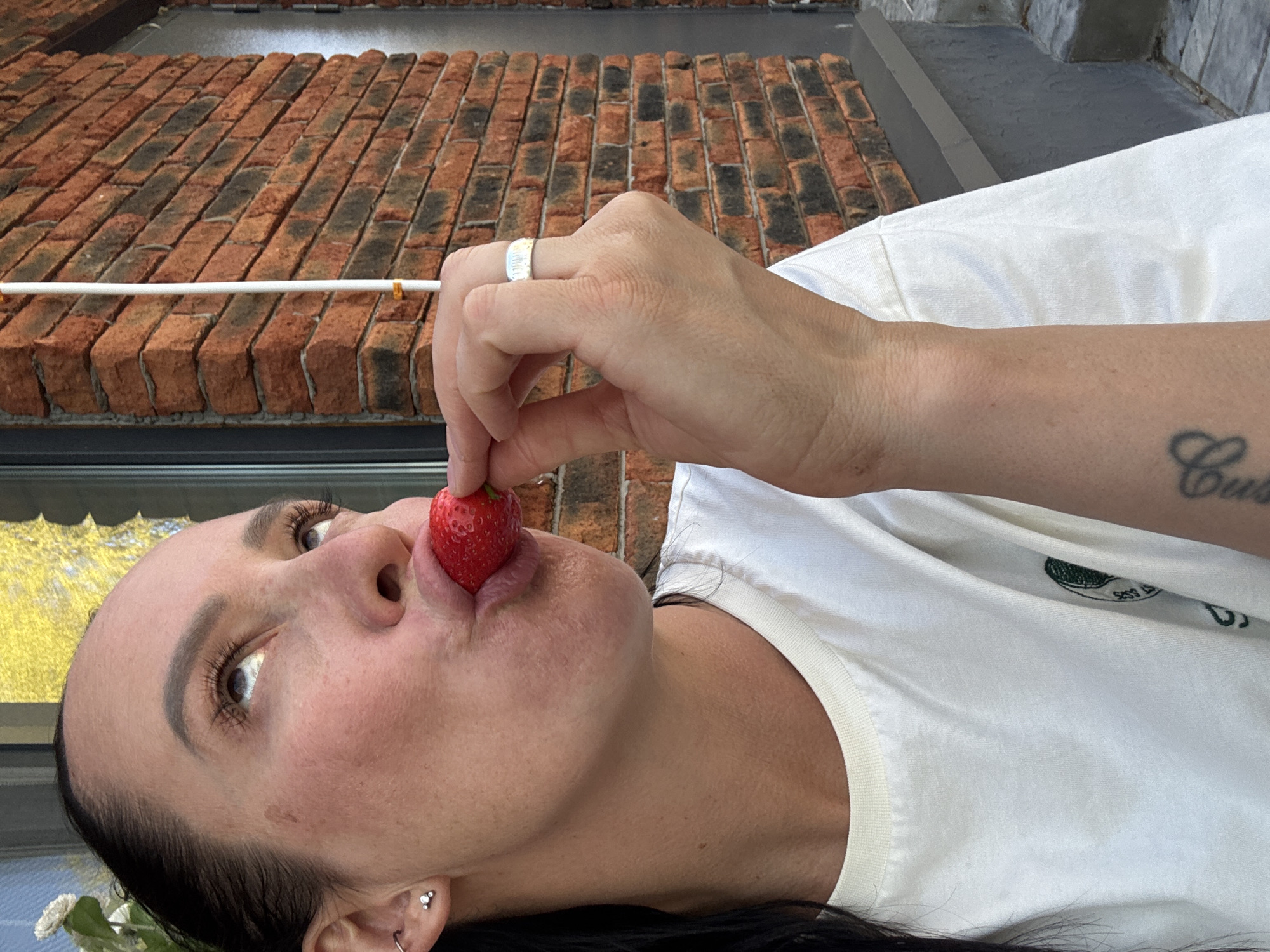 A woman is holding a strawberry to her lips. She has dark hair and is wearing a white t-shirt. She has piercings in her ear and a tattoo on her arm. She is wearing a ring on her finger. The background includes a brick wall and what appears to be an awning. The location is likely Chaikovsky, Russia, based on the prompt.