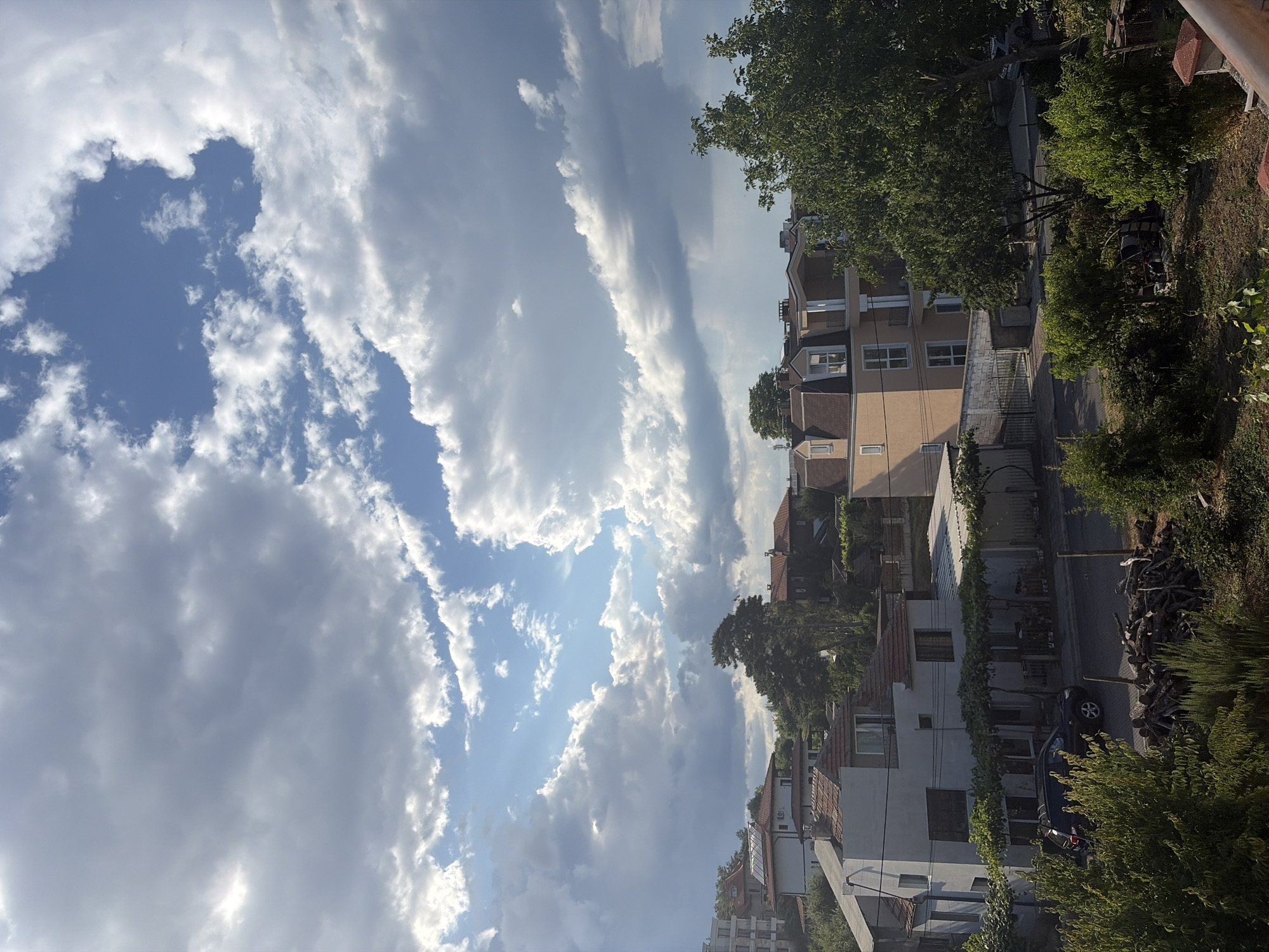 The image depicts a view of the city of Obzor, Bulgaria, from an elevated perspective. The scene is dominated by a dramatic sky filled with a mix of white and dark gray clouds, with patches of bright blue sky visible in between. Sunlight breaks through the clouds in some areas, casting a warm light on the landscape below.
In the foreground and midground, a residential area is visible. Several buildings with red-tiled roofs and light-colored facades are scattered amongst lush green trees. Some of the buildings appear to be multi-story houses or apartments. One prominent building in the midground has a beige facade with a mansard roof and multiple windows. A dark-colored SUV is parked on a paved street in front of one of the houses. A pile of cut logs is stacked on the ground near the street. The vegetation is dense, with trees and bushes covering much of the ground.
The overall impression is one of a semi-urban environment with a strong natural presence, likely captured during a time of changing weather, given the cloud formations. The lighting suggests it might be late afternoon or early morning, with the sun at a relatively low angle.
