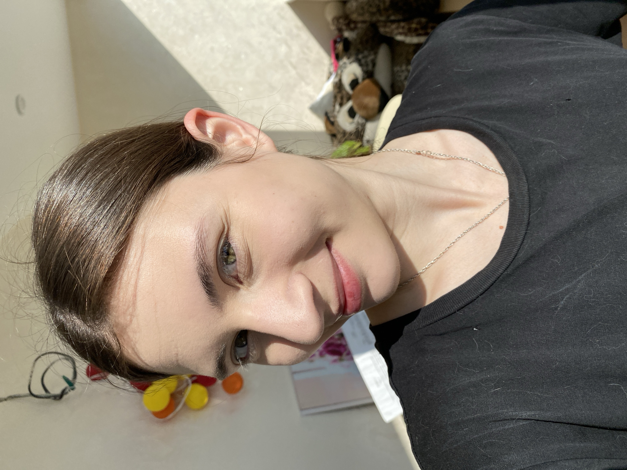 A woman with fair skin, light brown hair pulled back, and green eyes is facing the camera with a slight smile. She is wearing a black t-shirt and a silver necklace. Her head is tilted to the left. The background includes a window with bright sunlight streaming in. A toy or stuffed animal is partially visible near the window. The table she is on has multiple items, some of which appear to be toys. There is no indication this picture was taken in Votkinsk, Russia.