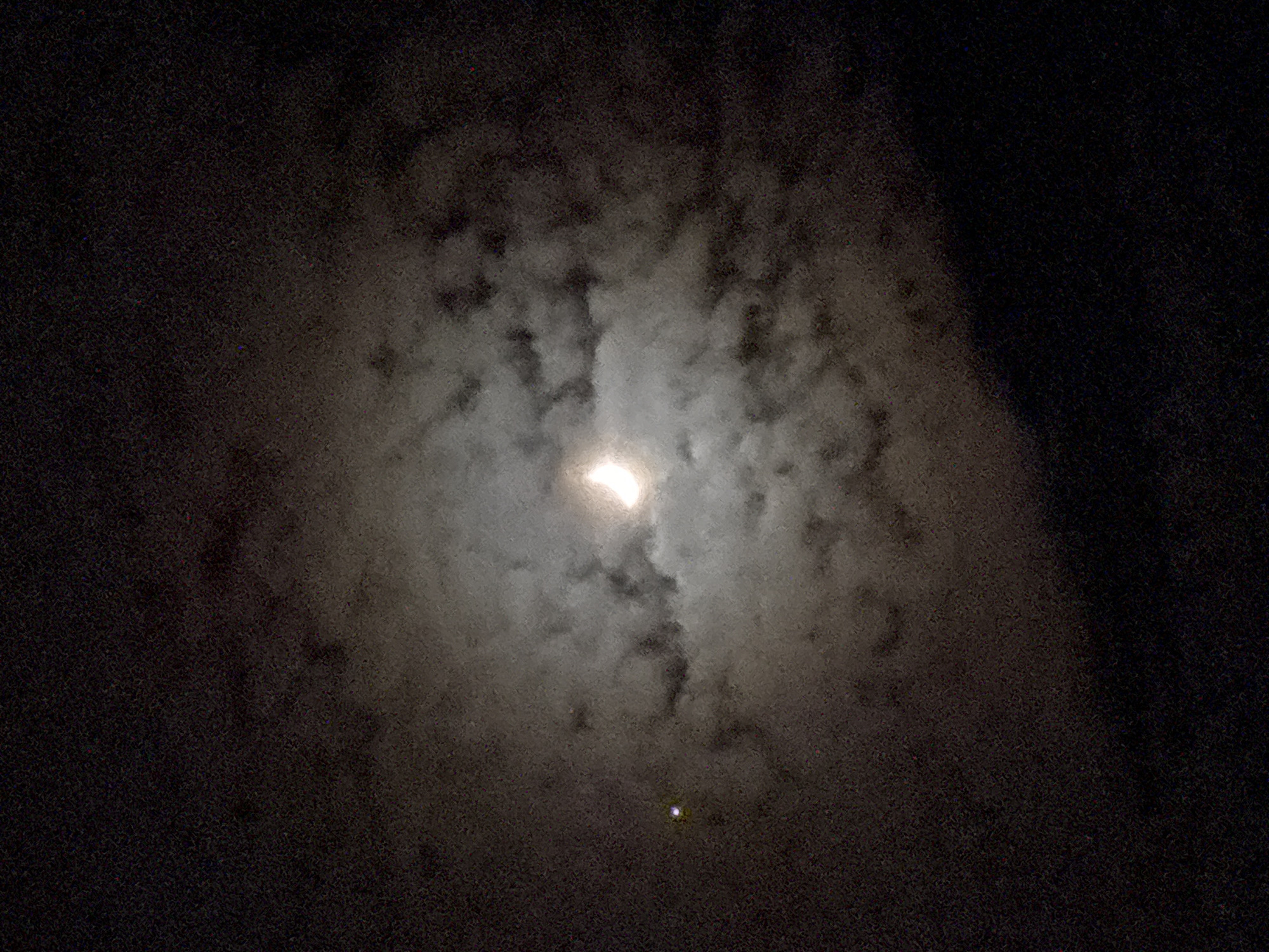 The image captures a night sky dominated by thick clouds. A very bright, diffuse light source, most likely the moon, is visible slightly above the center, glowing intensely through the clouds and backlighting them. The clouds appear as swirling patterns of light gray and white, contrasting against the darker, almost black areas of the sky where the clouds are denser or absent. Below the main light source, a smaller, distinct, bright white or yellowish-green dot is present, suggesting a star or a planet.
The time of day is night. The weather is cloudy, with the clouds sufficiently dense to obscure the distinct shape of the moon but allowing its light to diffuse widely. The scene suggests a celestial observation in a dark environment. The entire image has a grainy texture, especially noticeable in the darker portions of the sky, which is typical of low-light photography. There are no people, man-made objects, or specific location cues visible within the frame. No text is visible in the image.