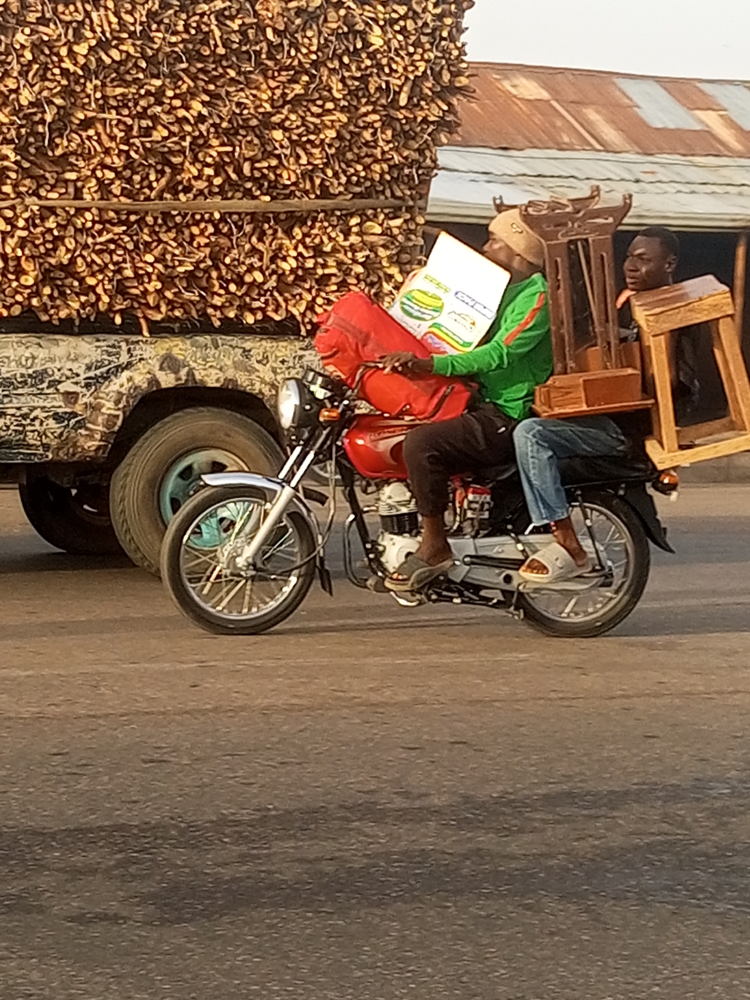 In Tumfure, Nigeria, a red motorcycle carries two individuals and various items. The driver, wearing a beige hat, green long-sleeved shirt, and brown pants, operates the motorcycle while holding a white box with colorful illustrations. Behind him, the passenger, clad in dark clothing, balances a disassembled wooden chair. A red bag rests on the motorcycle's front, near the handlebars. To the left, a truck piled high with bundled sticks stands parked. The background features buildings with corrugated metal roofs. The scene unfolds on a paved road.