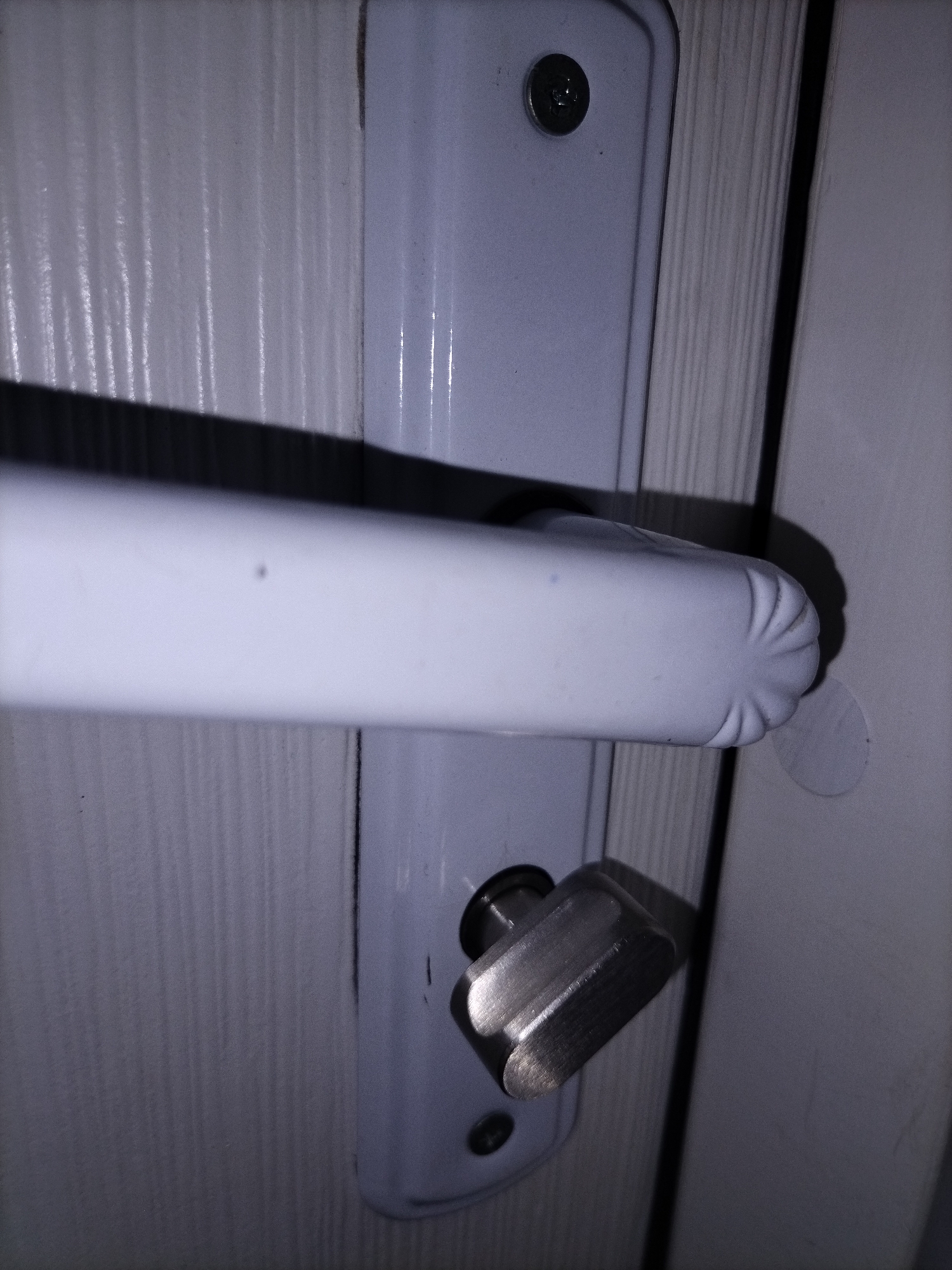 An extreme close-up shows a white door handle and lock mechanism against a white wooden door. The door handle is horizontal and elongated, with a ribbed end on the right. Below the handle is the lock mechanism, which appears to be made of brushed metal. The lock has a small knob that can be turned. The door has vertical wood grain visible. The lighting suggests the image was taken indoors, possibly in a room.