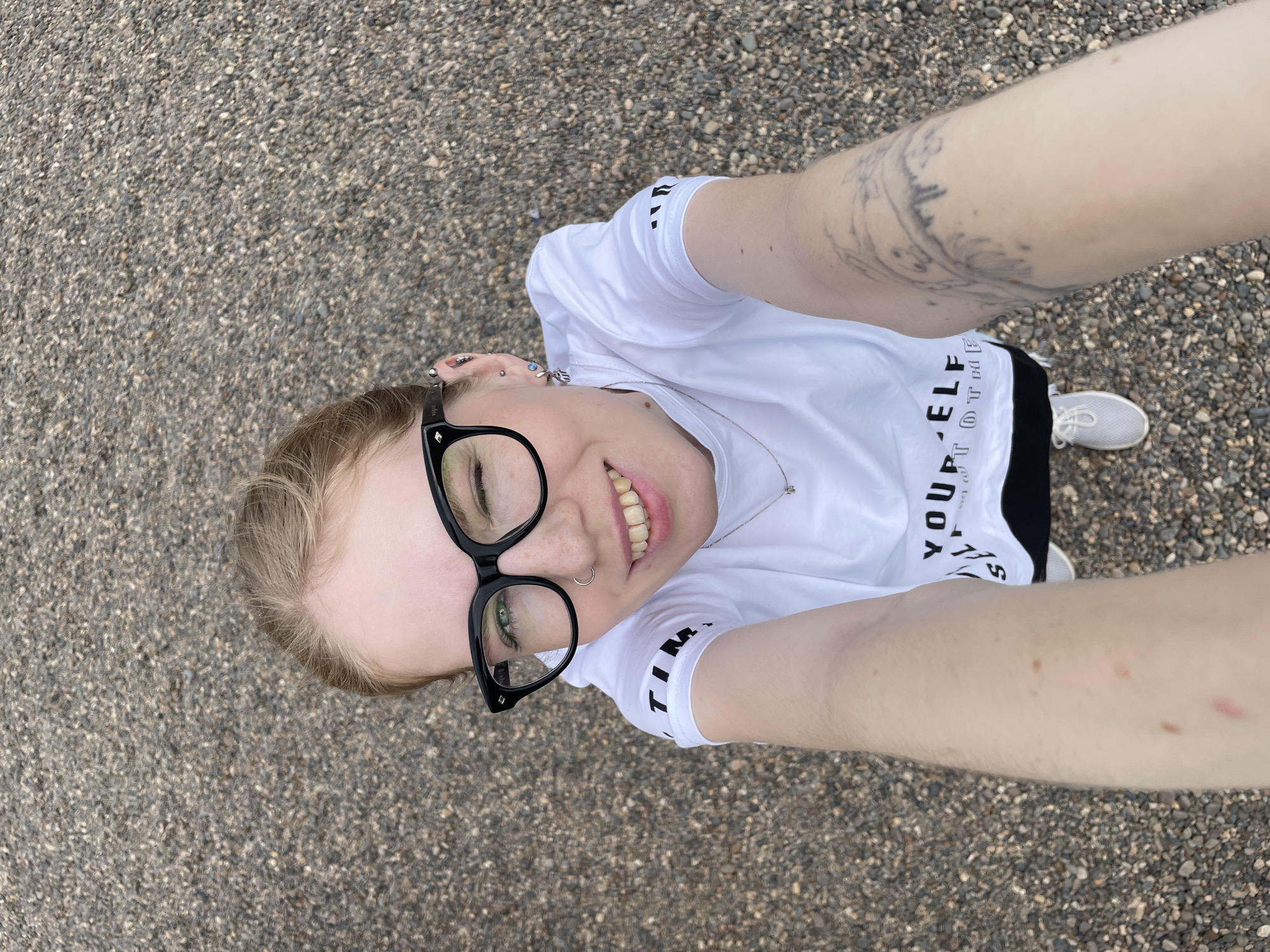 A light-skinned individual is standing on a gravel surface, likely a sidewalk or road, holding a camera or phone at arm's length to take a selfie from a high-angle perspective. The individual is wearing black framed glasses, a white t-shirt with text on the front ("YOUR SELF, NOT OTHERS"), a silver necklace, and a nose ring. Tattoos are visible on the individual's right arm. Only the top of the individual's white shoe is visible. The gravel surface comprises small, light-colored pebbles. The photo's location is unknown; there's no evidence to suggest the individual is in Izhevsk, Russia.
