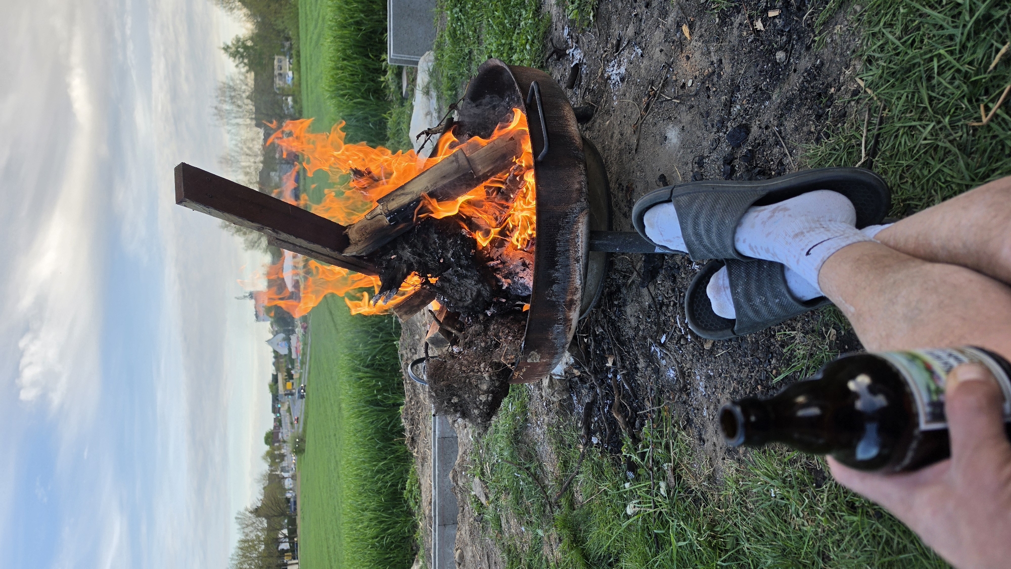 The image, rotated 90 degrees counter-clockwise, depicts a person relaxing by a burning fire pit outdoors. In the foreground, the lower legs and feet of a person are visible, wearing dark grey slide sandals over white socks, one of which clearly shows a black Nike swoosh logo. The socks appear soiled or stained. The person's left hand holds a dark brown glass bottle, likely a beer bottle, though its label is blurred and unreadable.
A round, metal fire pit dominates the central part of the image, filled with a vibrant bonfire. Large, bright orange and yellow flames engulf pieces of wood, producing smoke and glowing embers. The ground directly around the fire pit is a mix of dark soil, ash, and small debris, gradually transitioning into lush green grass.
In the background, beyond the immediate area, more green fields, scattered trees, and distant buildings, possibly houses, are visible. A road with faint lights or vehicles can be discerned in the far distance. The sky above is partly cloudy with patches of light blue and white clouds, indicating either late afternoon or early evening. The overall atmosphere suggests a casual and relaxed outdoor activity.