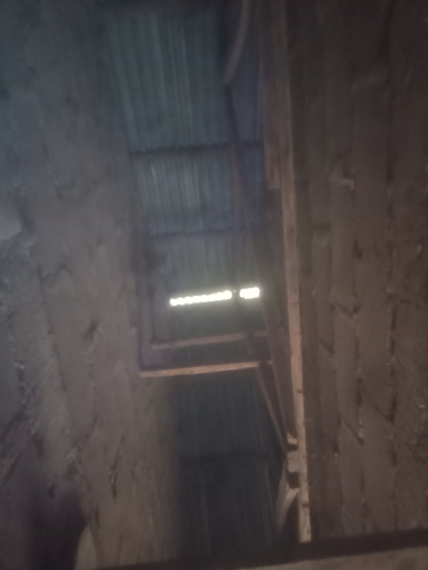 The image shows an interior view of what appears to be a construction site or an unfinished building. The perspective is looking upwards into a shaft or an opening in the ceiling. The walls on either side seem to be made of rough, unplastered brick or concrete. The ceiling itself is constructed with what looks like wooden planks or corrugated metal, with gaps allowing light to pass through. There are visible wooden beams supporting the ceiling structure. The lighting is dim, with a bright rectangular light source visible through an opening in the ceiling, suggesting natural light from above. The scene is devoid of any people or obvious objects of interest, conveying a sense of raw construction and an empty space. The location is unknown.