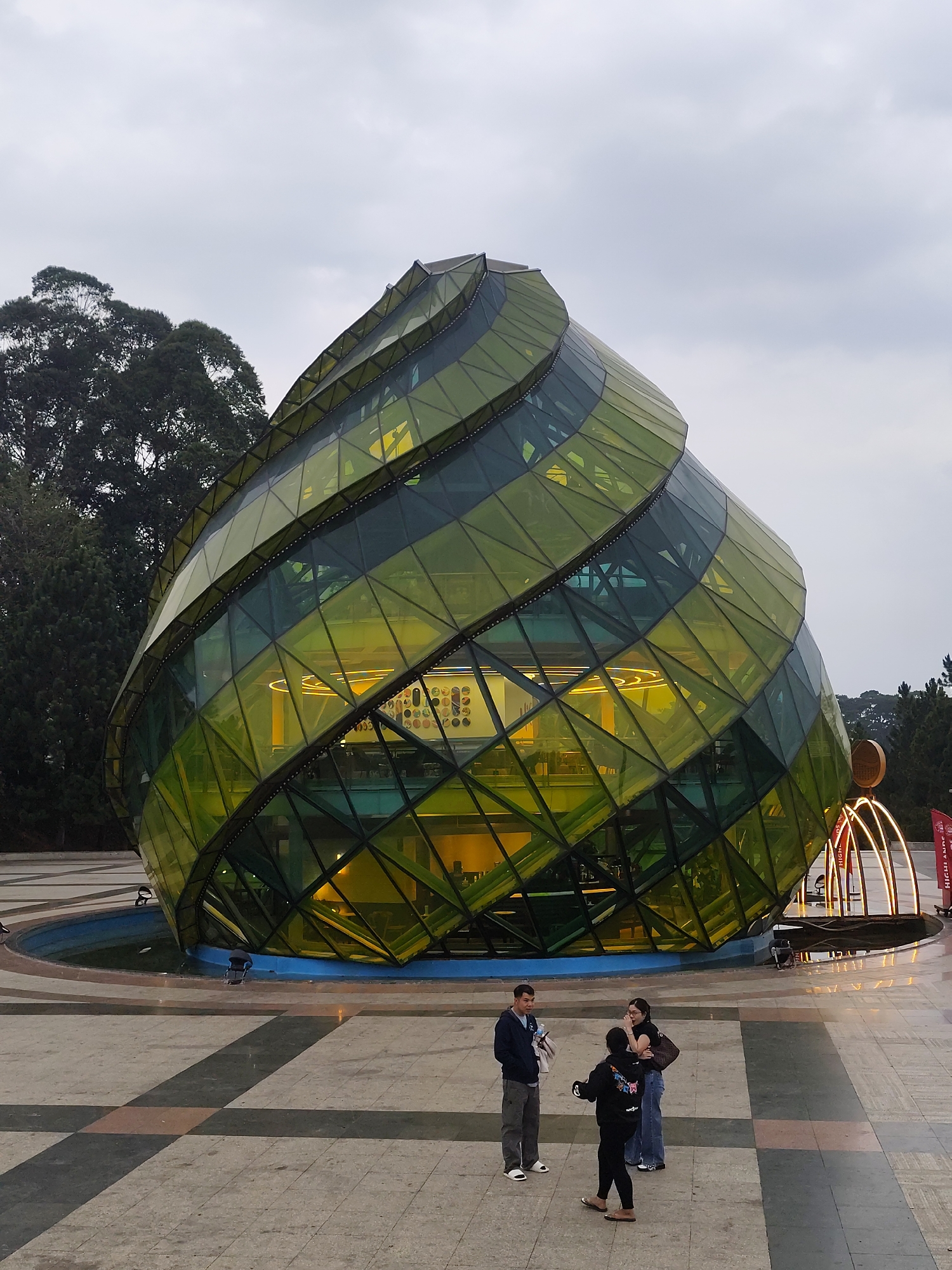 Here is a detailed description of the image:
The image shows a unique architectural structure in Da Lat, Vietnam, specifically at Lam Vien Square. This distinctive building resembles a giant artichoke or pine cone, constructed with green and yellow-tinted glass panels arranged in a spiraling pattern around a frame. The structure appears to be a building, with visible interiors, including what looks like a counter, lighting, and wall decorations inside. The exterior is illuminated with internal lighting.
The building sits on a tiled or paved plaza, surrounded by a circular water feature, though it appears dry. To the right of the main structure, there are illuminated curved arches, possibly an art installation, as well as flag poles.
In the foreground, a small group of three people is visible. A man stands in a navy-colored jacket, light gray pants, and white slides. He holds an object in his hands. A woman is standing next to him in a dark hoodie, black pants, and sandals. Another woman stands to the side in a light-colored jean jacket and jeans. Their posture suggests they are engaged in a conversation or interaction.
The background contains a large tree with dense foliage, and the sky is overcast, suggesting a cloudy day. The overall scene appears to be a tourist attraction or public space.