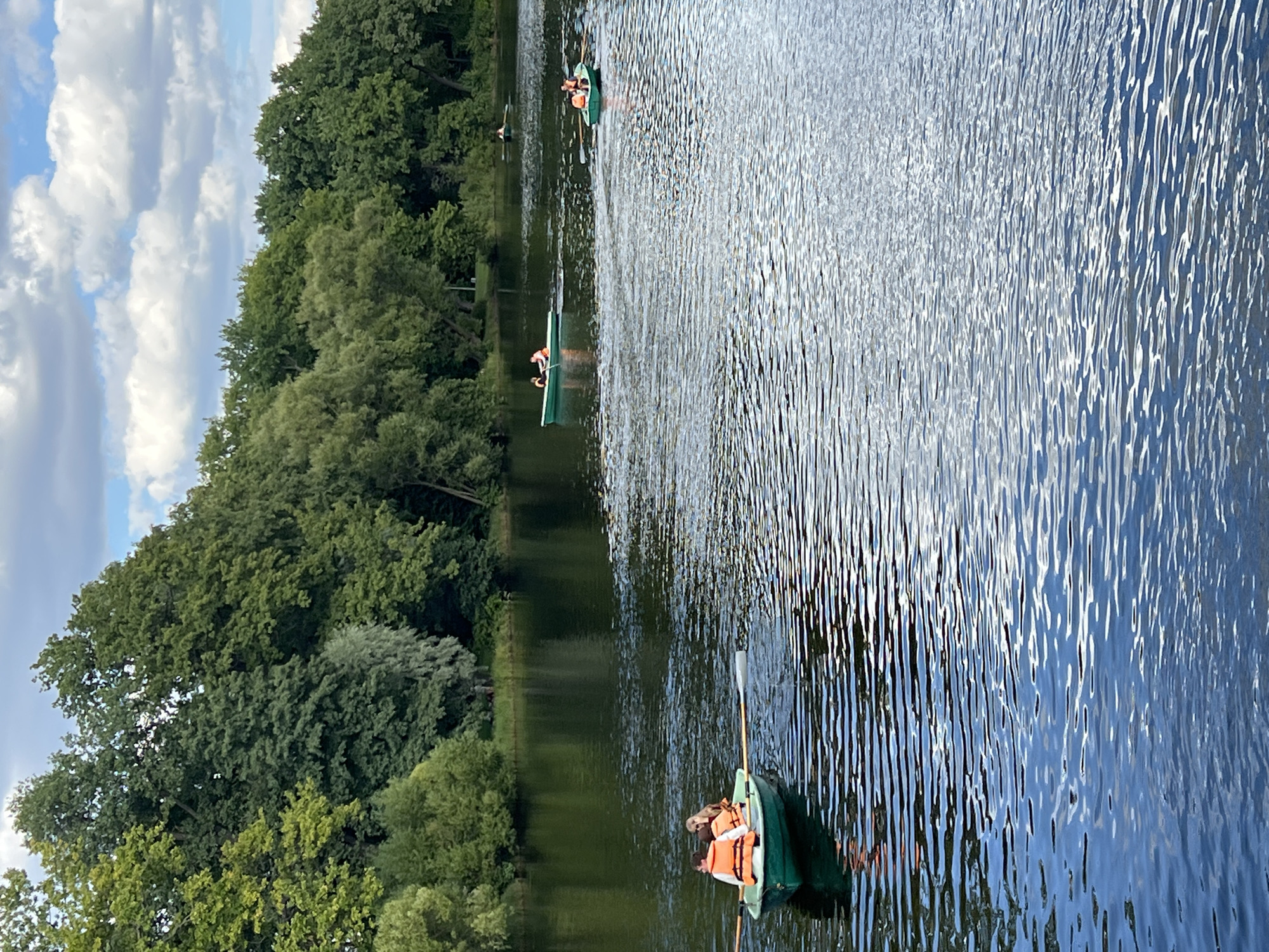 The image depicts a scenic view of a tranquil lake bordered by lush green trees, under a sky with scattered white clouds. Several rowboats are visible on the water. In the foreground, a green rowboat carries two individuals wearing orange life vests, one of whom is rowing. Further in the distance, two other green rowboats are also visible on the lake, with occupants wearing life vests. The water's surface is rippled, reflecting the surrounding greenery and the sky. The scene appears to be in a park or natural reserve, suggesting a leisurely outdoor activity. Given the mention of Saint Petersburg, Russia, this scene could be located in one of its many parks with bodies of water, such as the Pavlovsk Park or the Catherine Park, which are known for their scenic landscapes and boating activities.