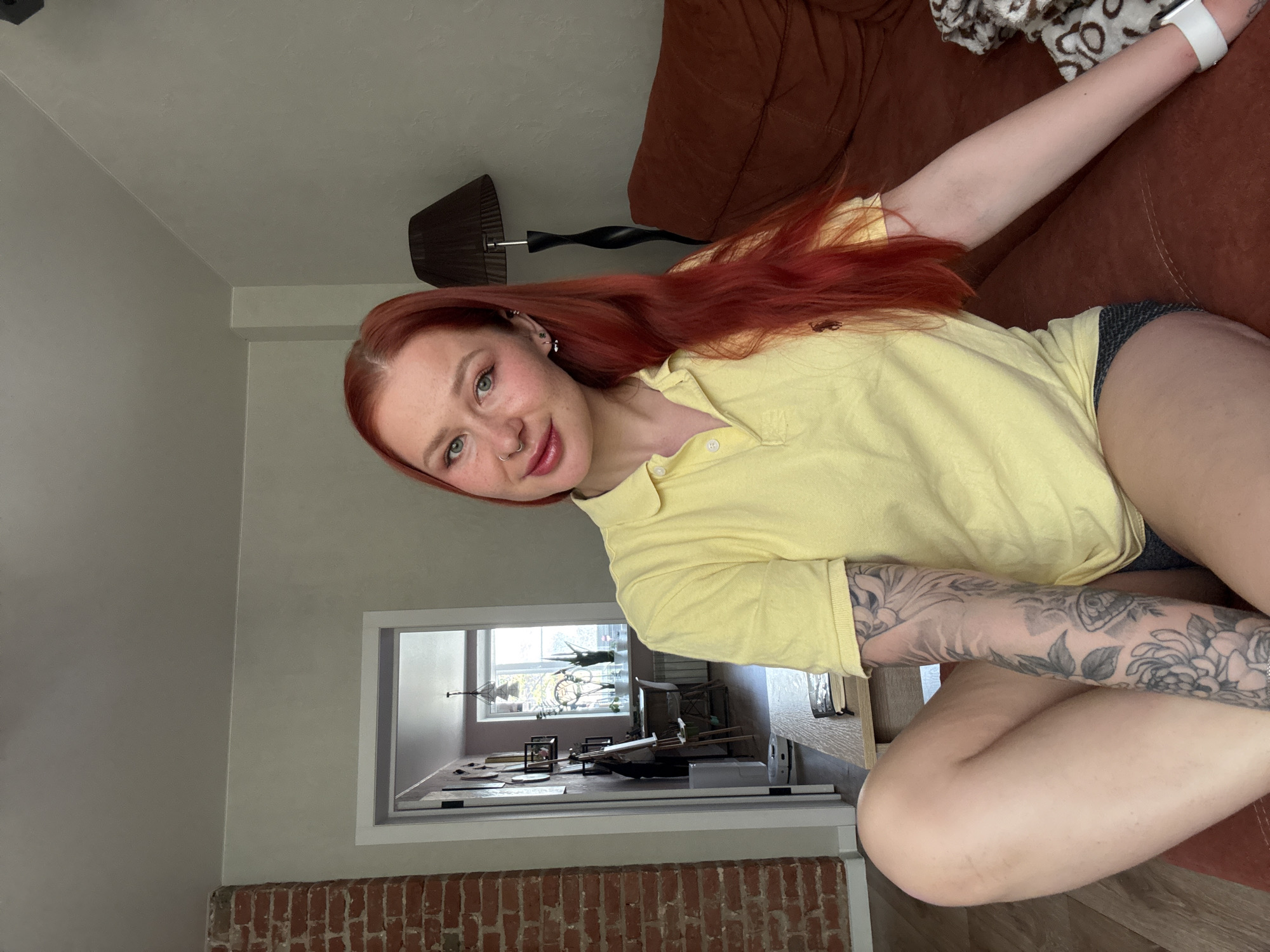 A woman with red hair and green eyes is sitting on a brown couch. She is wearing a yellow polo shirt and dark shorts. She has a tattoo on her left arm. A lamp is behind her on the couch. A doorway with a view of a room with various items can be seen in the background. Brick detail is on the wall near the doorway. The location is not specified in the image.