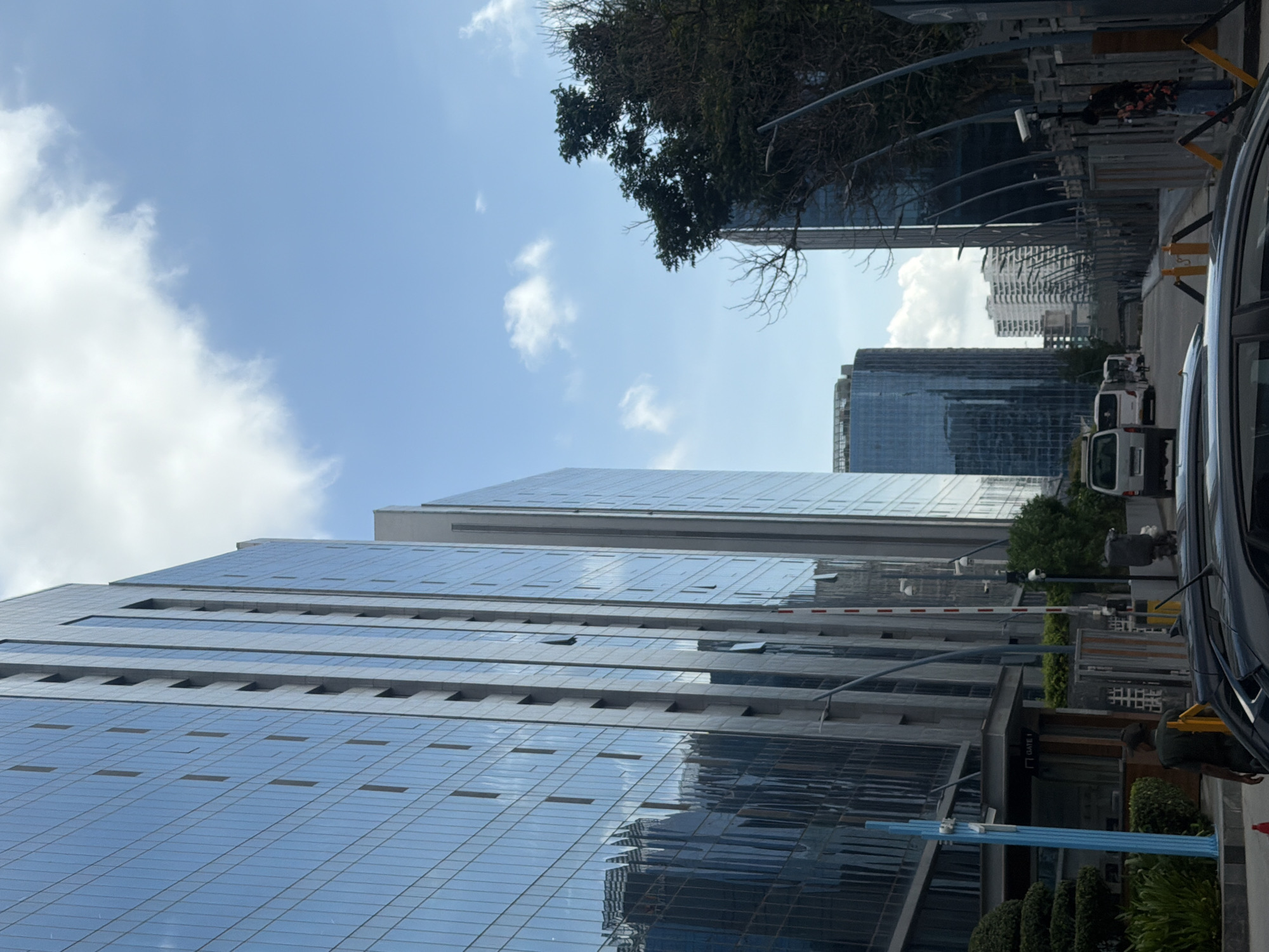 This image depicts a street scene in Hyderabad, India, likely in a modern business or IT district. Two tall, glass-faced buildings dominate the left side of the frame, reflecting the sky and clouds. These buildings appear to be modern office complexes. In the mid-ground, a road runs between the buildings, with several vehicles, including a white van and other cars, visible. A few people are present, including one person walking away from the camera near a security booth with a "GATE 1" sign. Another person is standing near a fence on the right side of the road, looking at their phone. The right side of the image features another tall building, partially obscured by a tree with bare branches. The sky is blue with scattered white clouds. The overall impression is of a bustling urban environment with contemporary architecture.