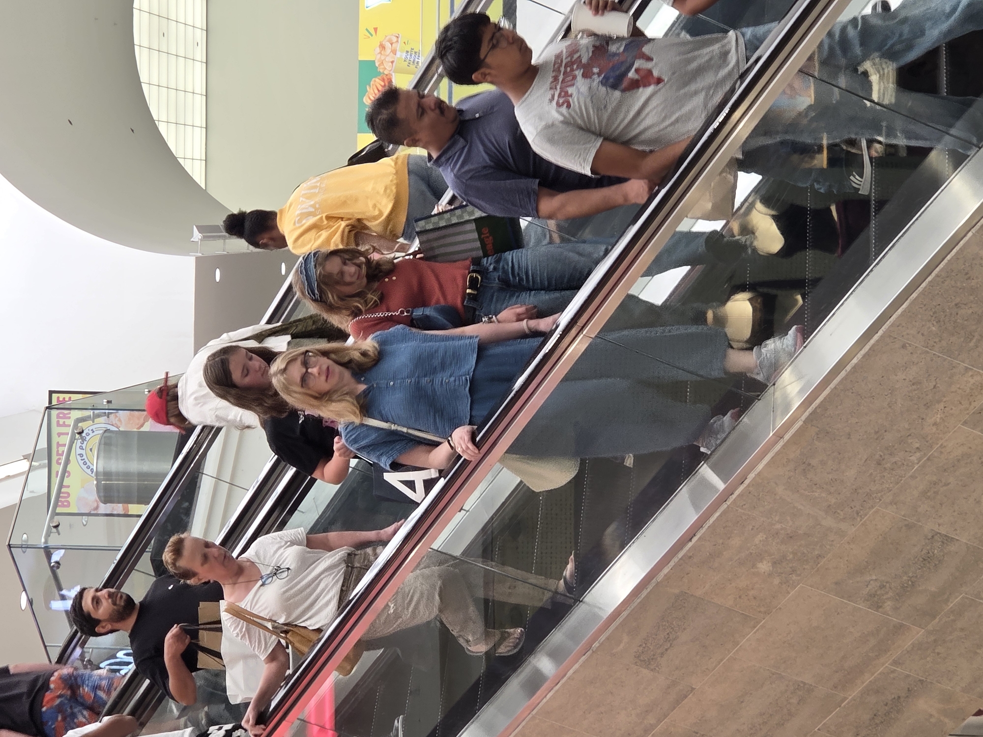 The image shows a group of people on an escalator in a public indoor space, possibly a shopping mall. The escalator has a polished metal railing and a marble-like tiled side. The lighting appears to be artificial, coming from overhead. In the foreground, a woman wearing a blue denim-like outfit is ascending the escalator. To her left and slightly behind her, another woman in a white t-shirt and patterned pants is also on the escalator. Above them, a man in a black t-shirt is holding shopping bags. Further up the escalator, there are other individuals. In the background, a bright yellow sign is visible, possibly an advertisement. The scene appears to be taking place during the daytime. The individuals are dressed in casual clothing.