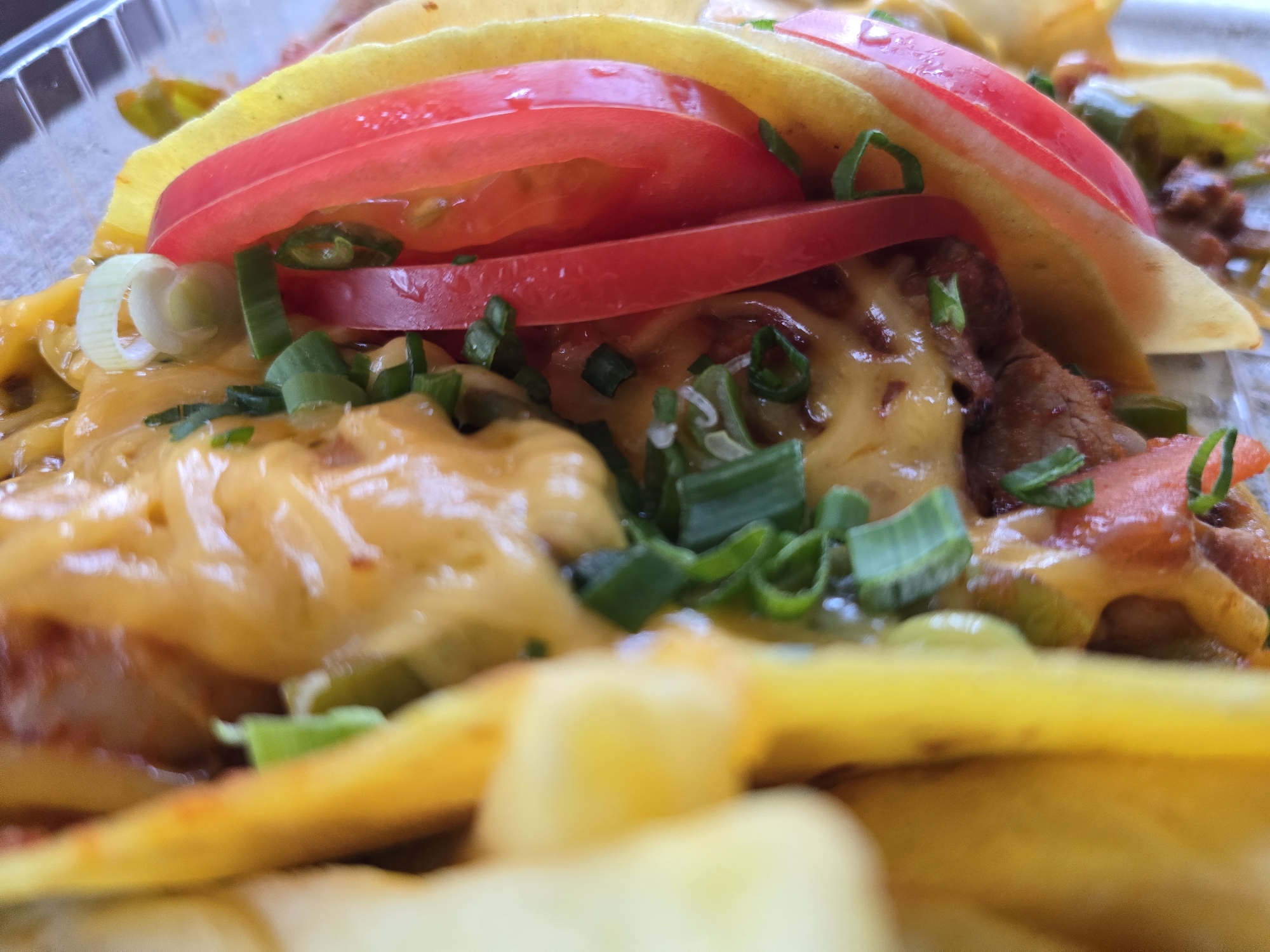 This close-up shot presents a dish of tacos, likely Mexican-style, featuring soft yellow tortillas filled with melted cheese, meat, possibly beef, sliced tomatoes, and chopped green onions. The food is contained within a clear plastic container, suggesting it is takeout or pre-packaged. The location is not discernible from the image.