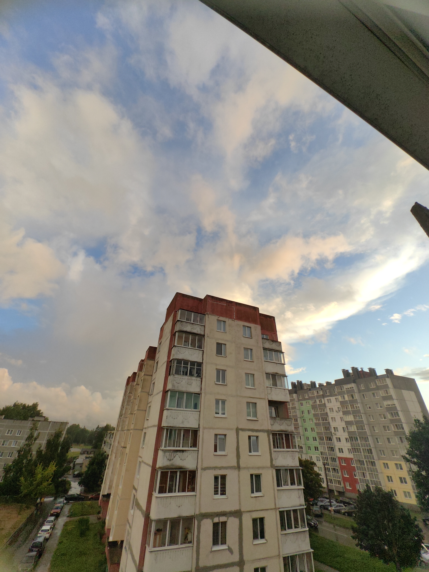 The image displays a residential area in Fanipal, Belarus, under a cloudy sky. Several multi-story apartment buildings dominate the scene, characterized by their concrete facades and numerous windows. One building in the foreground, with a red-trimmed roof, shows balconies and a mix of window types. To the right, another apartment building is visible, featuring a colorful facade with sections painted in green, red, and yellow. The ground level of the area shows a street with parked cars and patches of green grass and trees. The perspective suggests the photo was taken from a higher vantage point, possibly from another apartment building.