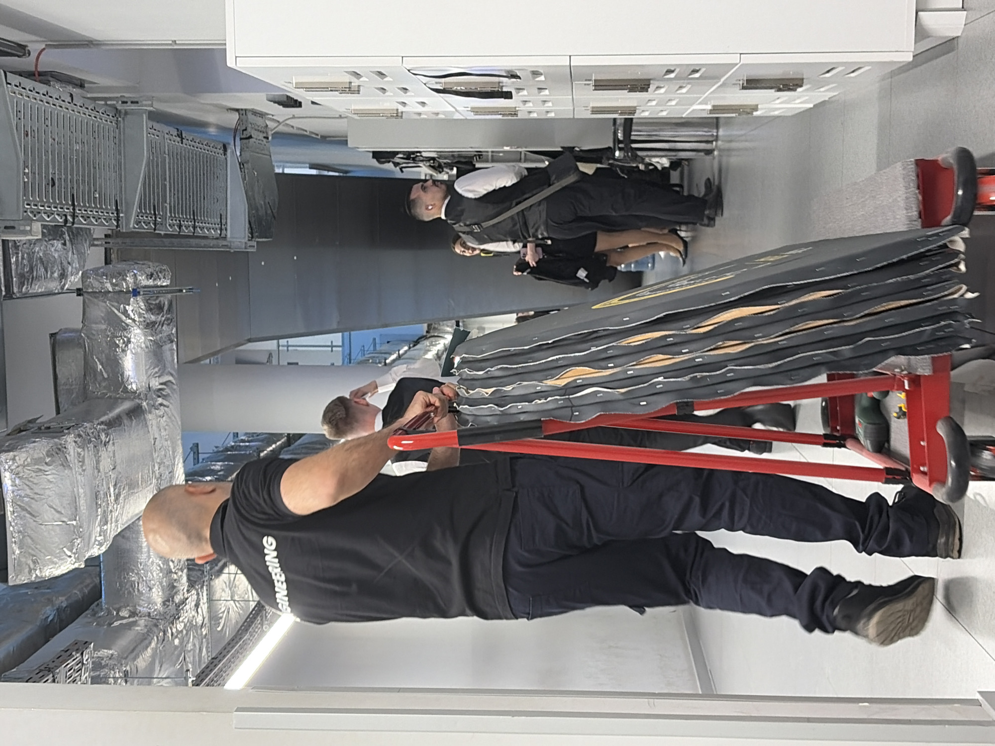 The image depicts an indoor scene, likely a hallway or corridor, with a focus on a man wearing a black polo shirt with "ENGINEERING" in white lettering on the back. He is standing next to a red cart carrying folded, dark-colored panels with metallic accents. The cart also has a green drill on it. Behind him, another man in a white shirt and dark vest stands near a row of white lockers. Further in the background, a woman in a black skirt and white top is visible. The environment includes exposed ductwork with silver insulation on the ceiling and white walls. The floor is light gray tile. There is no indication of the city of Sofia, Bulgaria, within the visual information provided.