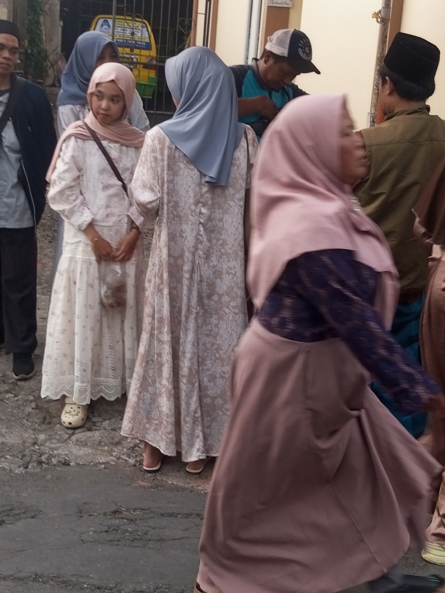 In Pandansari, Indonesia, on a grey, overcast day, a group of individuals stands on a paved street. The scene is dominated by several women in traditional Islamic attire. In the foreground, a woman in a light pink hijab and a matching long dress is seen from the side, her right arm extended. Behind her, another woman in a pale blue hijab and a white floral patterned dress holds a small plastic bag. To the left, a young woman in a pink hijab and a similar white floral dress is looking towards the viewer, also holding a plastic bag. Further back, a man wearing a blue shirt and a baseball cap is visible, along with another man in a black cap and a olive green vest. A portion of a yellow vehicle, possibly a bemo or public transport vehicle, is visible in the background, partially obscured by a dark metal fence. The ground is a rough, textured asphalt.