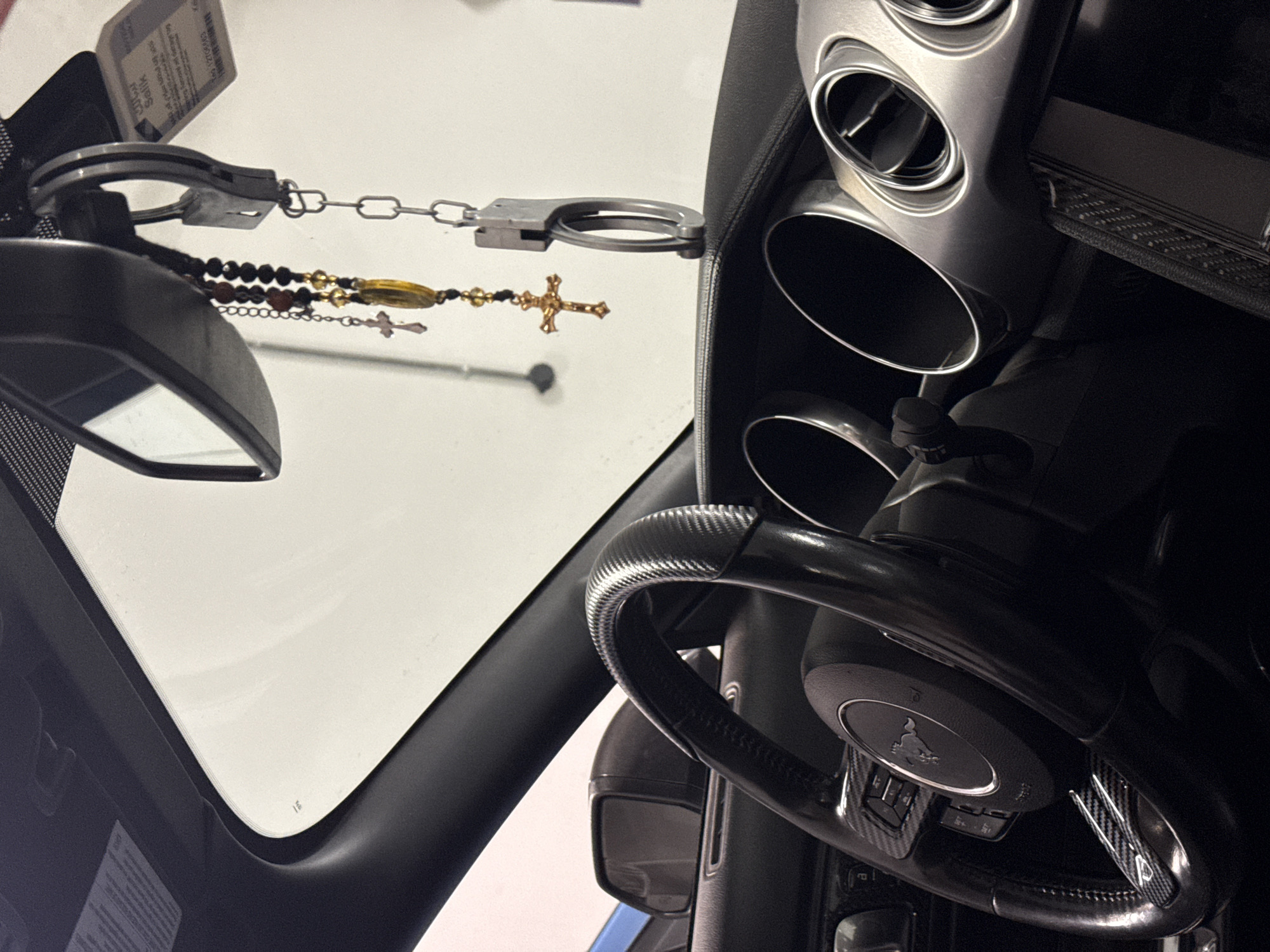 The photograph shows an interior view of a vehicle, specifically the driver's side. A steering wheel with carbon fiber accents is visible, along with dashboard elements, air vents, and a rearview mirror. Handcuffs and a rosary are suspended from the rearview mirror. The outside view, reflected by the windscreen, shows a white surface and suggests an indoor setting. There is no discernible information within the image to indicate a location in Dubai, United Arab Emirates, or any other specific geographical location.
