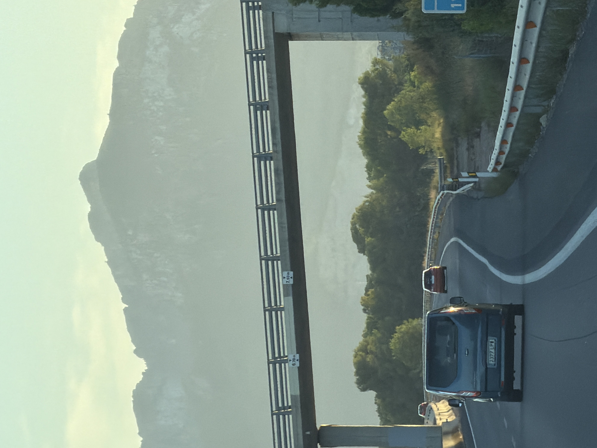 A highway overpass is visible in the foreground, with a mountain range in the hazy background. The overpass has two height restriction signs, one indicating 6.70m and the other 7.50m. A dark-colored van is driving on the highway in the right lane, with a license plate reading "2377 LPW". In the distance, two other cars, one red and one orange, are also on the highway. Trees and vegetation line the sides of the road, and a guardrail is present. The sky is a pale blue, with some light clouds. The scene appears to be a rural or suburban area in Mont-roig del Camp, Spain, during daylight.