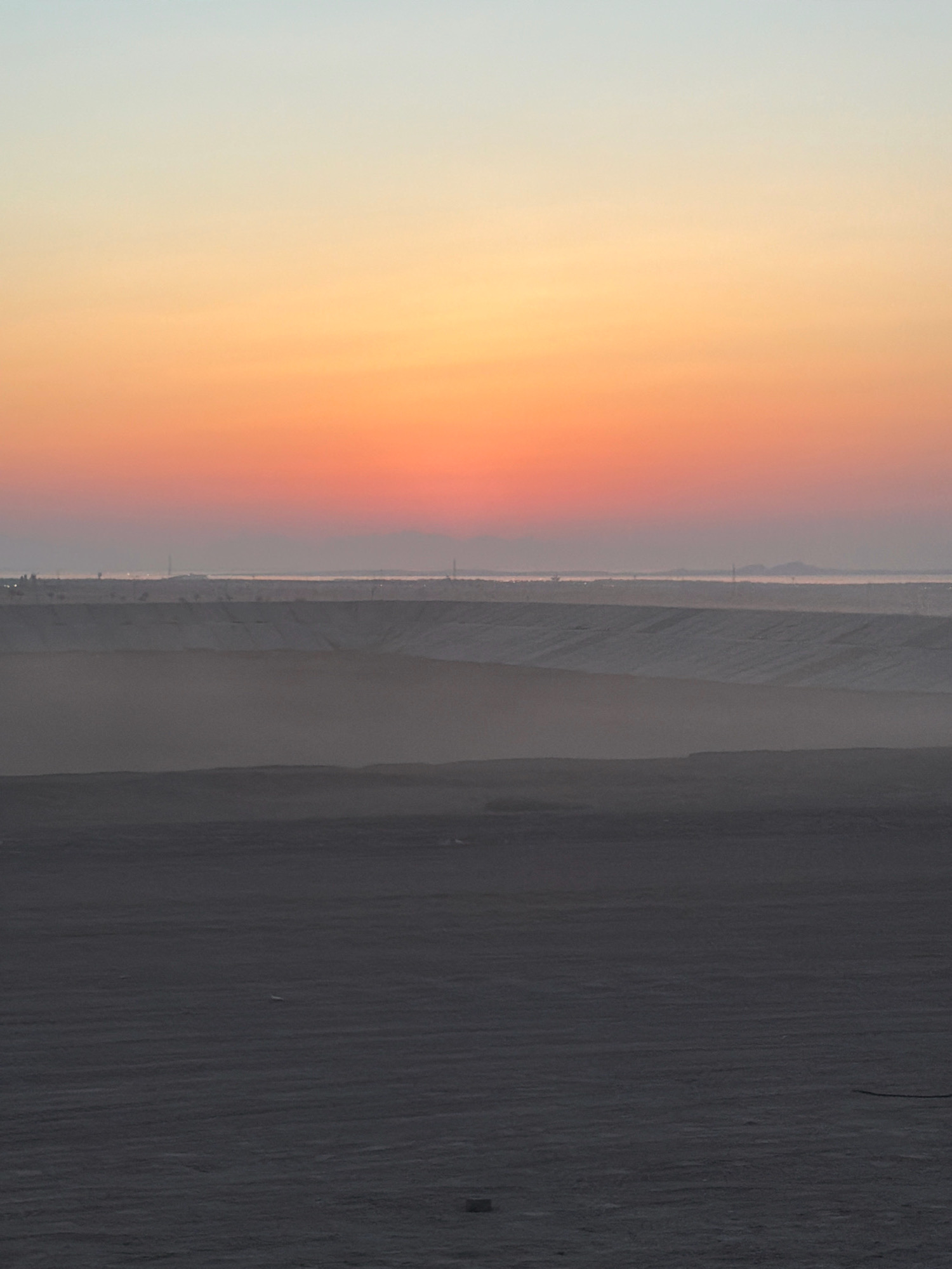 The image depicts a vast desert landscape at dusk, with the sky transitioning through soft hues of blue, orange, and pink. In the foreground, the ground is dark and textured, with subtle horizontal lines suggesting the surface of sand dunes or a prepared area. Layers of progressively lighter, undulating terrain recede into the distance, shrouded in a hazy atmosphere. Far in the background, a distant coastline is faintly visible, marked by structures and lights, hinting at an urban or industrial presence. The overall scene is serene and expansive, characteristic of the desert outskirts of Sharm El Sheikh, Egypt, likely captured during the twilight hours. There are no people or animals visible in the image.