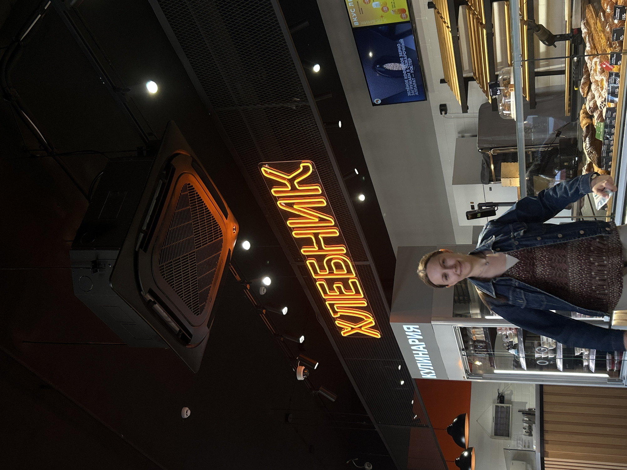In a brightly lit bakery or cafe in Saint Petersburg, Russia, a young woman with blonde hair, wearing a denim jacket over a patterned top, smiles towards the camera. She is standing in front of a display case filled with baked goods, including loaves of bread. Above her, a neon sign with Cyrillic text reads "ХЛЕБНИК" (Khlebnik), which means "baker" or "bread maker." To the right of this sign, another sign in Cyrillic reads "КУЛИНАРИЯ" (Kulinariya), indicating "culinary" or "delicatessen." A digital screen displays text in Russian, mentioning "green coffee beans" and a "roasting machine." The overall atmosphere suggests a bustling, well-stocked establishment. The lighting is bright, and the scene appears to be indoors, likely during the daytime given the interior illumination. The woman's expression is relaxed and friendly, suggesting she is either a customer or an employee enjoying the environment.