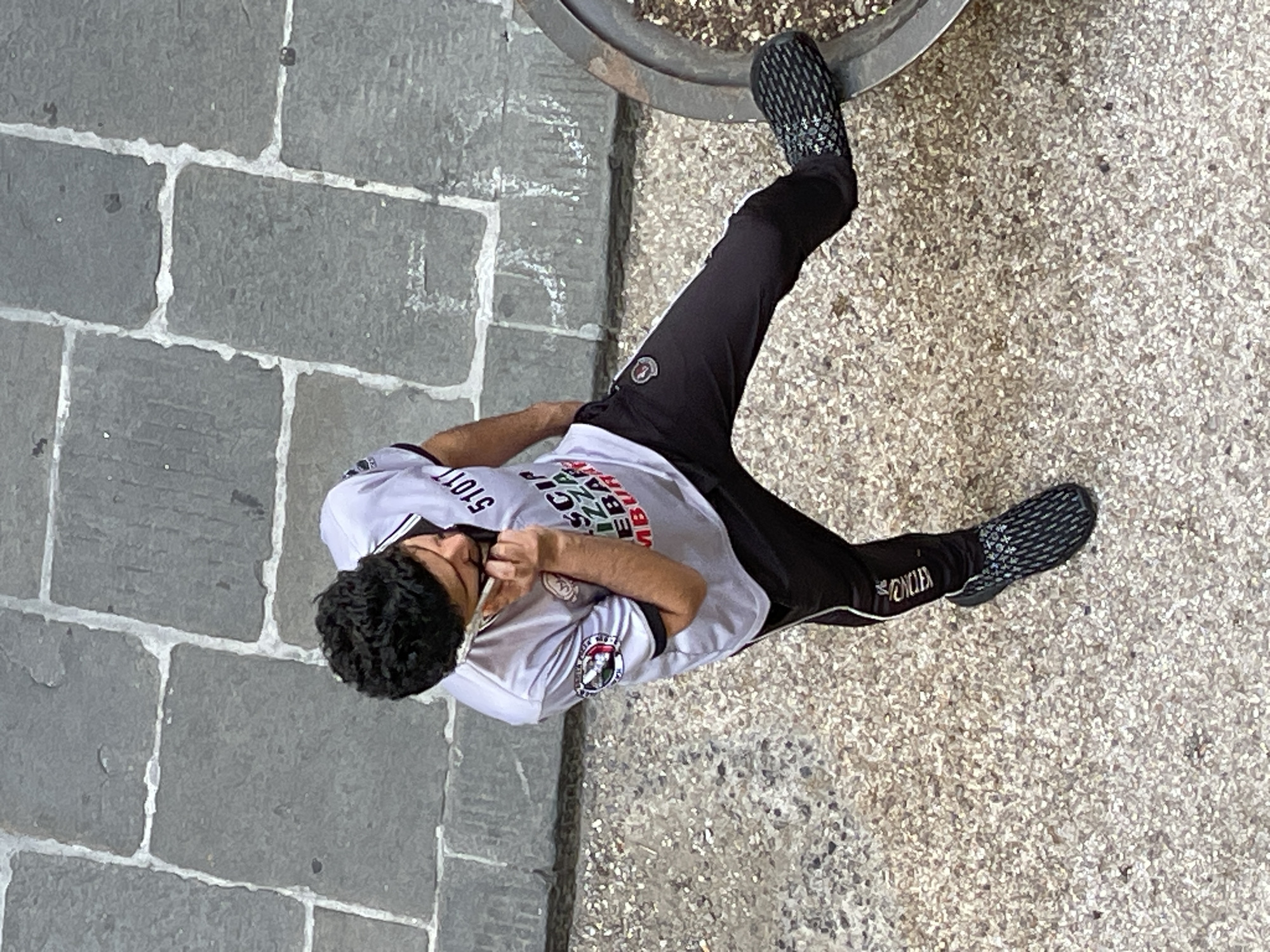An overhead medium shot captures a man speaking on a smartphone while standing on a cobblestone and gravel surface. The man, who appears to be of adult age, is wearing a light gray short-sleeved shirt with dark trim and black athletic pants with a white stripe down the side. The shirt features graphics, including the number "51011" and text that is partially obscured but appears to include "SCIP", "PIZZA", "EBA", and "MBUR". A circular patch with a crest is visible on the left sleeve. He is holding a white smartphone to his right ear and his right hand is raised towards his face. His left hand is in his pants pocket. He is wearing black athletic shoes with a patterned sole. His right leg is extended forward and slightly bent at the knee. To the right of the man, a portion of a large, dark-colored planter or basin is visible. The ground is composed of large, irregular gray cobblestones with white grout in the upper portion of the image, transitioning to a speckled gravel surface in the lower portion. The scene appears to be an outdoor urban environment.