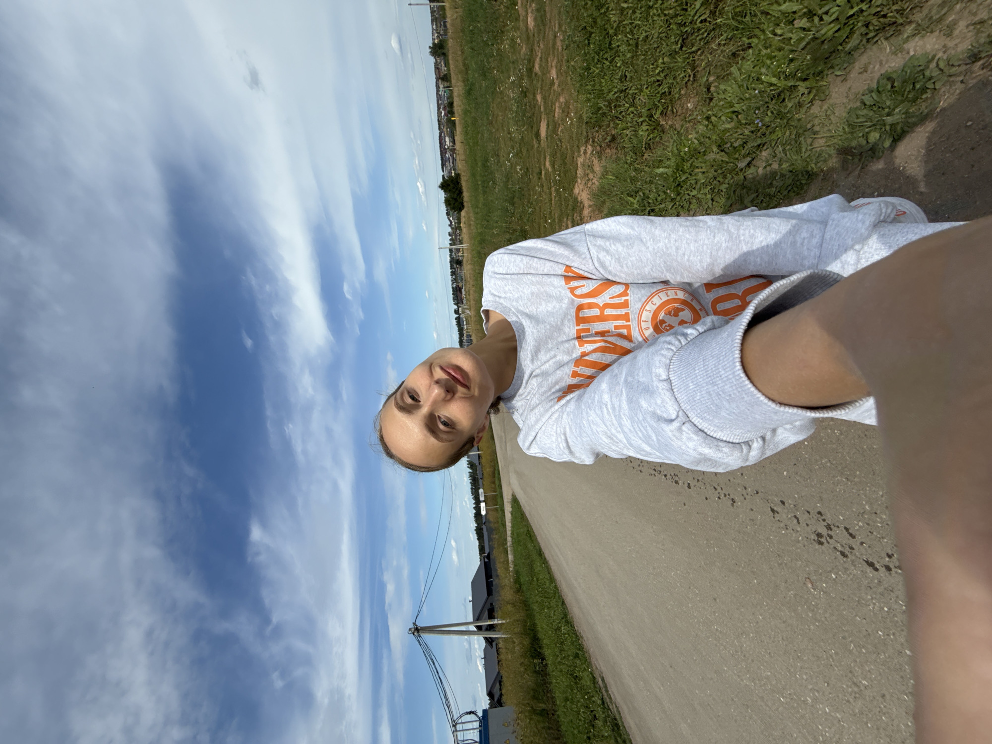 A woman with blonde hair pulled back smiles at the camera, holding the camera out to take a selfie. She is wearing a gray sweatshirt with orange text. She stands on a dirt road in what appears to be a rural area. The background shows a field of dry grass and some houses with dark roofs. There are power lines overhead and a blue sky with white clouds. The location is Podshivalovo, Russia.