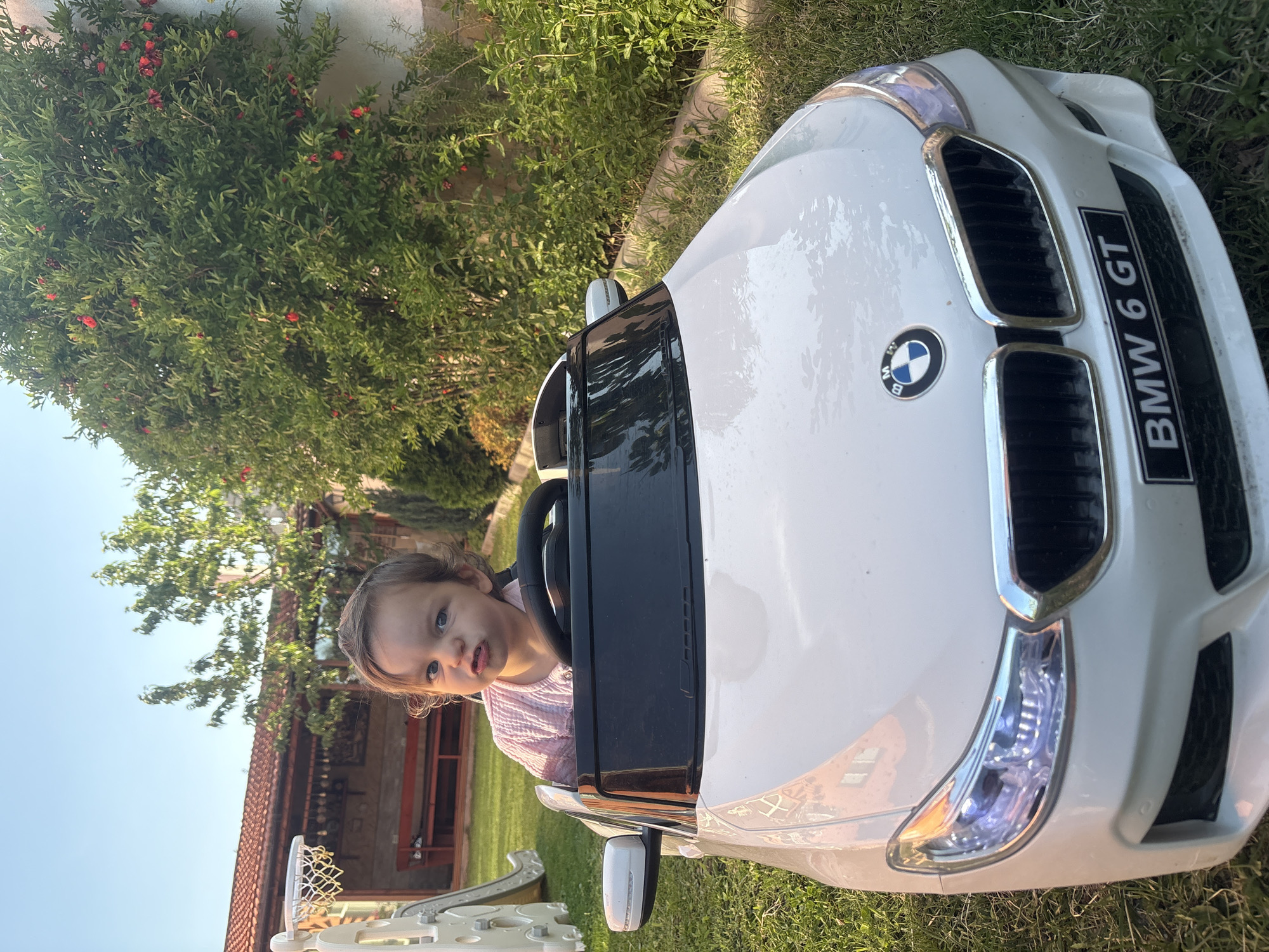 In a backyard setting in Strelcha, Bulgaria, a young child sits in a white toy BMW 6 GT. The child, with brown hair and a light complexion, wears a pink, textured shirt and makes a pouty facial expression. The toy car is parked on green grass, with a paved path visible behind it. In the background, a building with a red tile roof is partially visible, along with a basketball hoop and a playground slide. Lush green foliage, including a tree laden with small red fruits, surrounds the scene. The sky is clear and blue. The lighting suggests it is late afternoon.