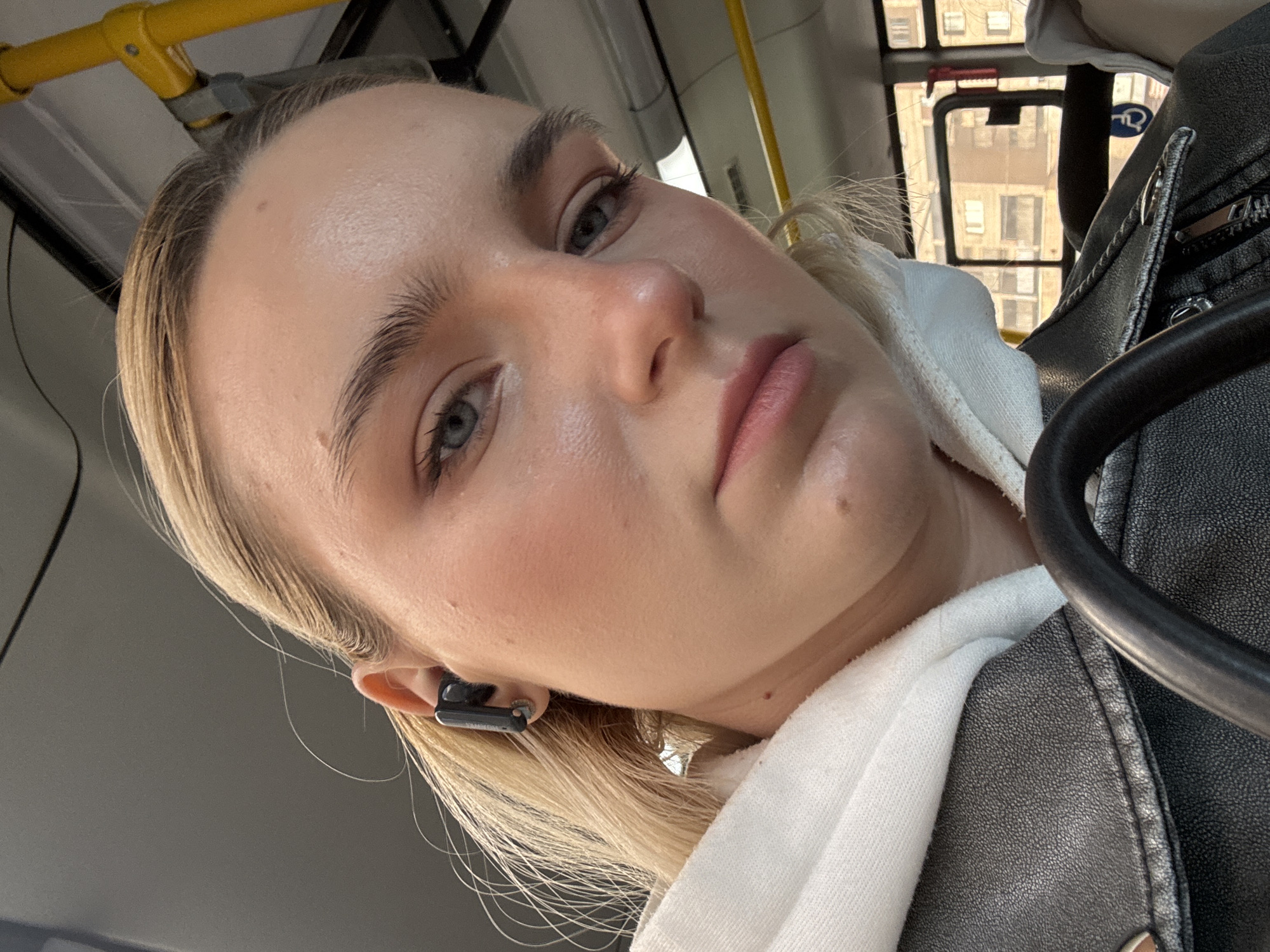A close-up, tilted selfie shows a fair-skinned person with blonde hair. They have light eyebrows and gray eyes. The subject is wearing a black earbud in their left ear, a white hooded sweatshirt, and a black leather jacket. The background suggests they are on a bus, with yellow handrails and a window showing buildings outside. The location is not discernable as Vsevolozhsk, Russia.