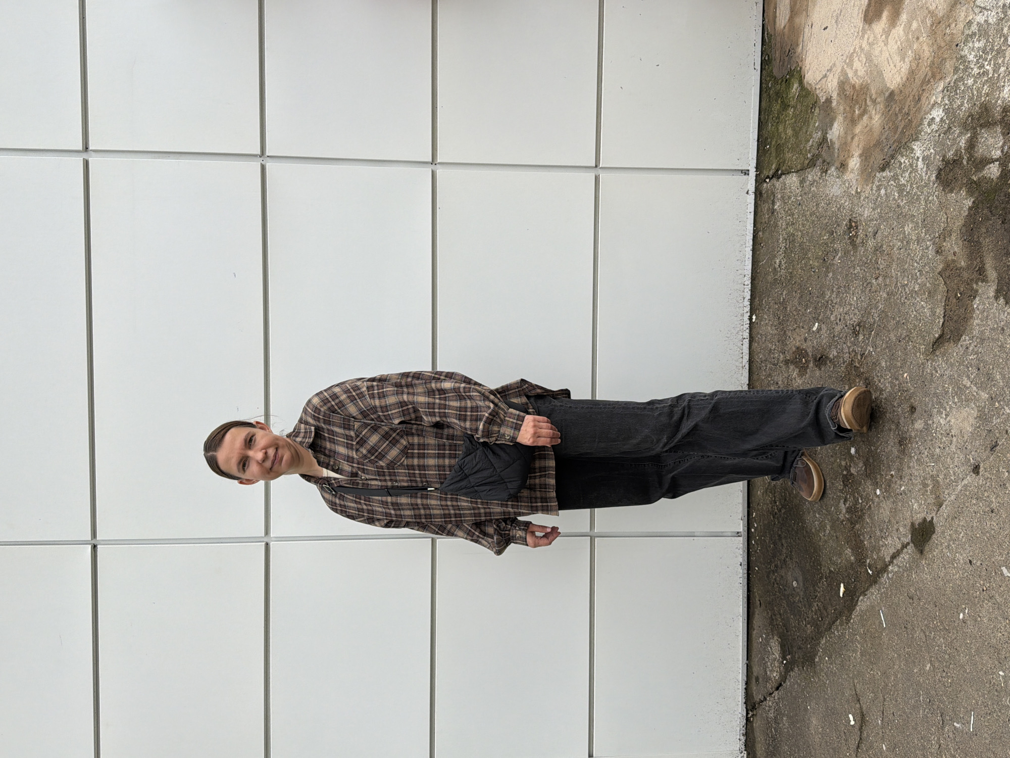 The image features a person, oriented vertically, standing in front of a white tiled wall. The person is wearing a brown and beige plaid shirt, dark denim jeans, and brown shoes. A small black quilted bag is slung across their chest. Their hair is pulled back, and they are looking towards the camera with a slight smile. The background consists of the uniform white tiles of the wall, which are divided into a grid pattern. To the right of the wall, there is a patch of weathered concrete with some green and brown discoloration, suggesting it's an outdoor setting. The lighting appears to be natural, potentially indicating daytime. The image is taken from a slightly low angle, looking up at the subject. There is no visible text in the image.