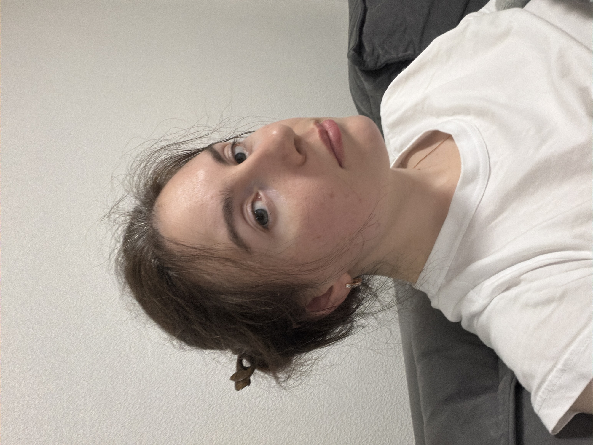 A woman with fair skin and light blue eyes is looking directly at the camera. She has brown hair pulled back in a low bun, with some strands framing her face. She is wearing a white t-shirt and is seated or reclining against a grey sofa or upholstered surface. The background is a plain white wall. The image is taken from a slightly elevated perspective, with the camera angled downwards. The lighting is soft and even. There is no indication of the location being Vsevolozhsk, Russia.