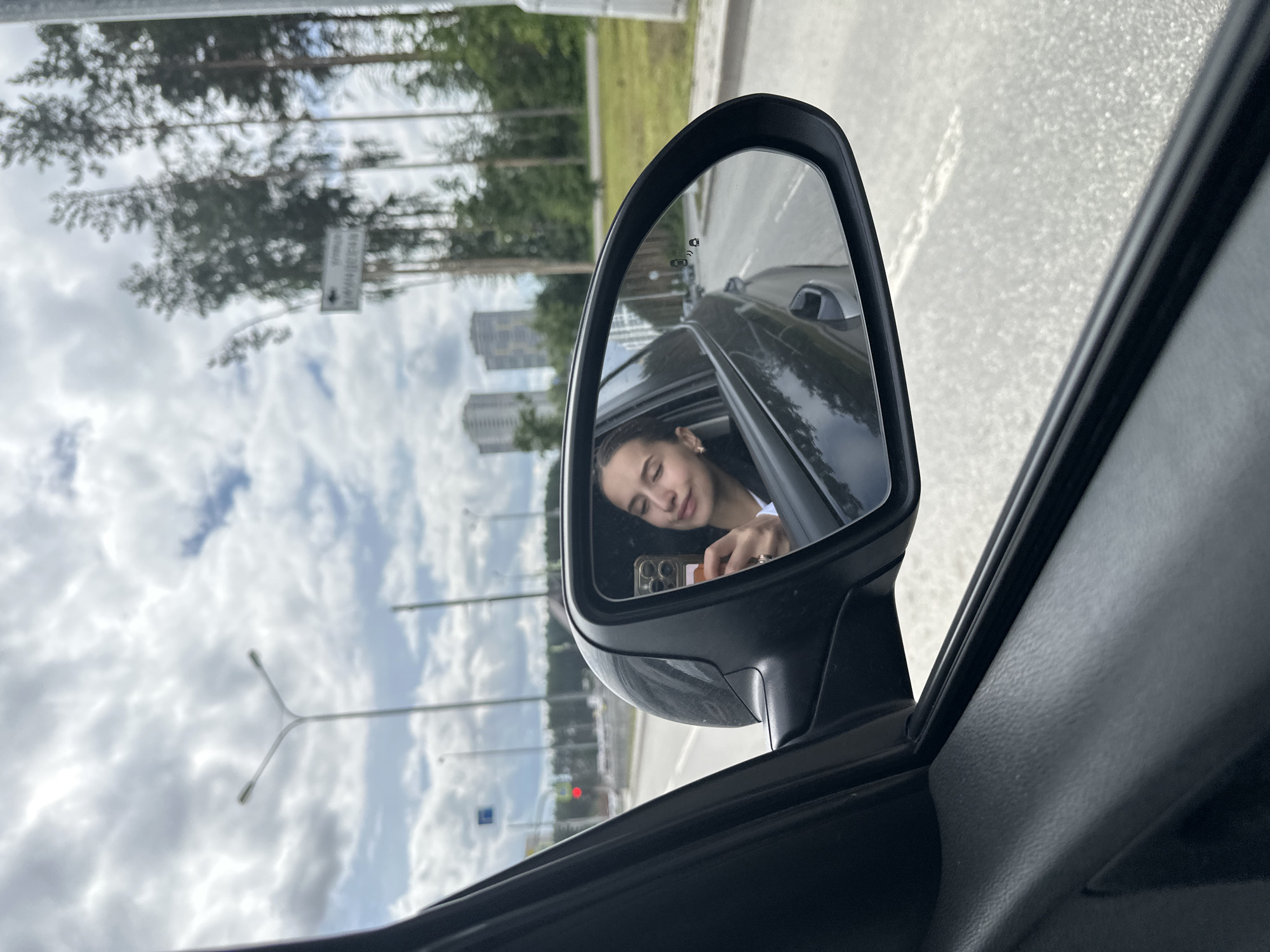 A young woman is reflected in the side-view mirror of a car, taking a selfie with her phone. She is looking at the phone with a slight smile, and her hair is pulled back in braids. The background in the mirror shows the road, trees, and buildings. In the foreground, the interior of the car is visible. The scene appears to be a suburban or highway area in Yekaterinburg, Russia, with characteristic tall trees and modern buildings visible. The sky is cloudy.