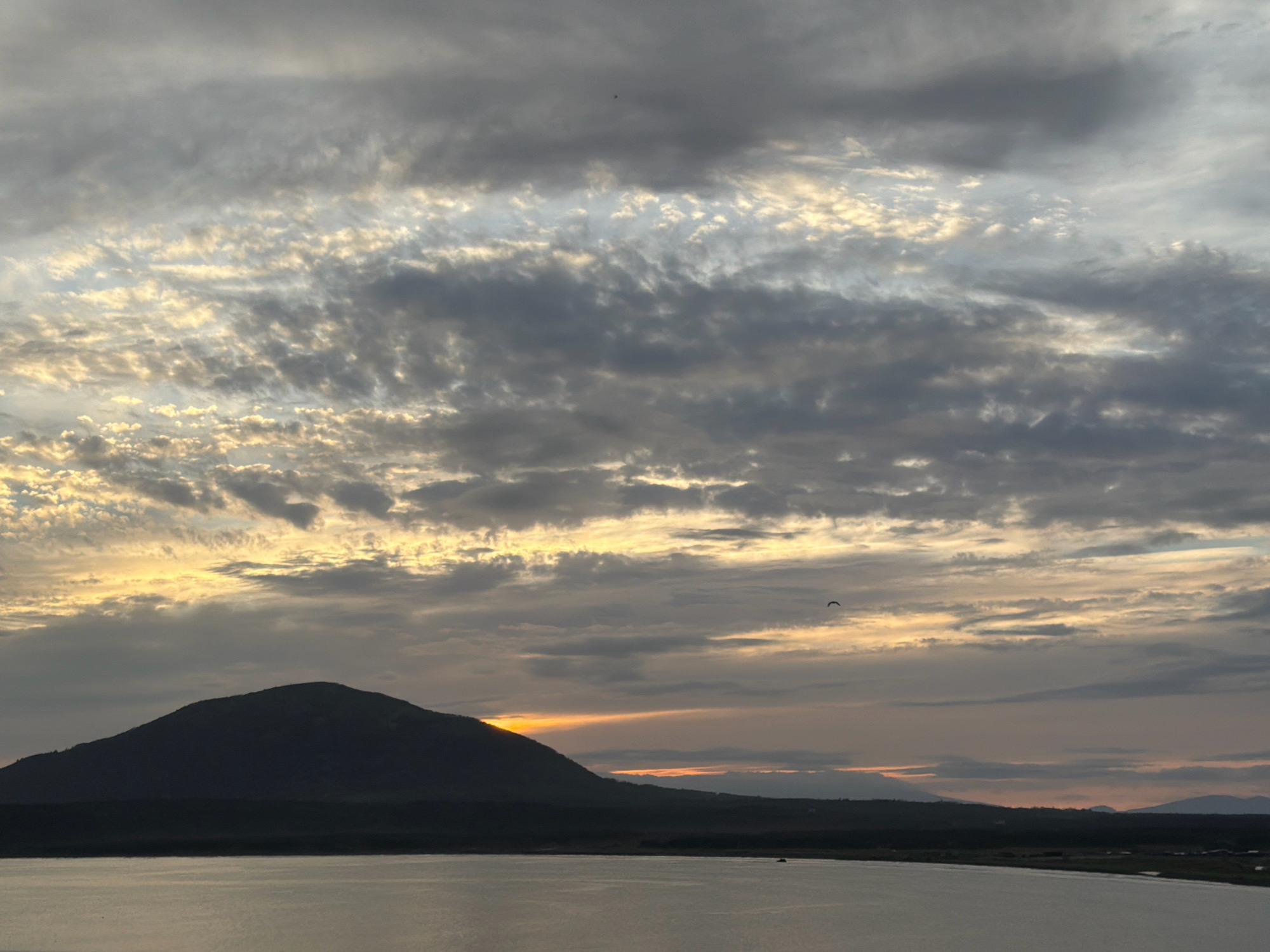 The image depicts a scenic vista of a body of water in the foreground, with a silhouetted mountain range occupying the middle ground. The sky is filled with dramatic cloud formations, illuminated by the warm hues of a sunset or sunrise, suggesting a coastal location. The faint outlines of distant hills and what appear to be structures or settlements can be observed along the horizon. One of the cropped images shows a single bird in flight against the textured clouds. The overall impression is one of natural beauty and tranquility. The location is identified as Yuzhno-Kurilsk, Russia.