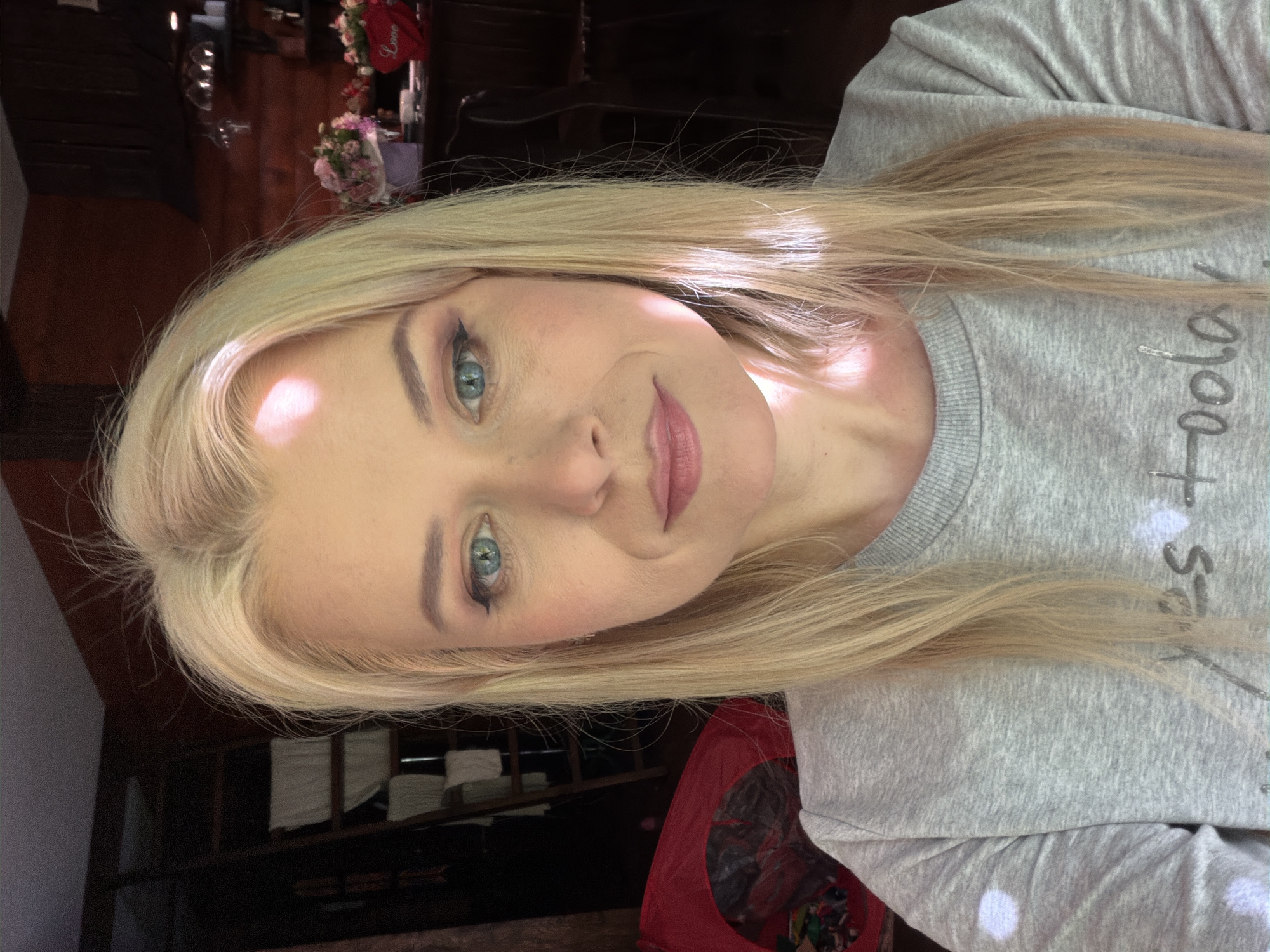 A close-up, head-and-shoulders selfie shows a blonde woman with blue eyes wearing a grey sweatshirt. She is indoors in what appears to be a rustic room with wood-paneled walls and a ladder in the background. Sunlight is streaming in from the right, casting bright highlights on her face and hair. On a counter in the background, there are two vases of flowers and a red heart-shaped decoration that says "Love." The woman's makeup is visible, including eyeliner and eyeshadow. The overall lighting suggests it is daytime. The location is specified as Sochi, Russia.