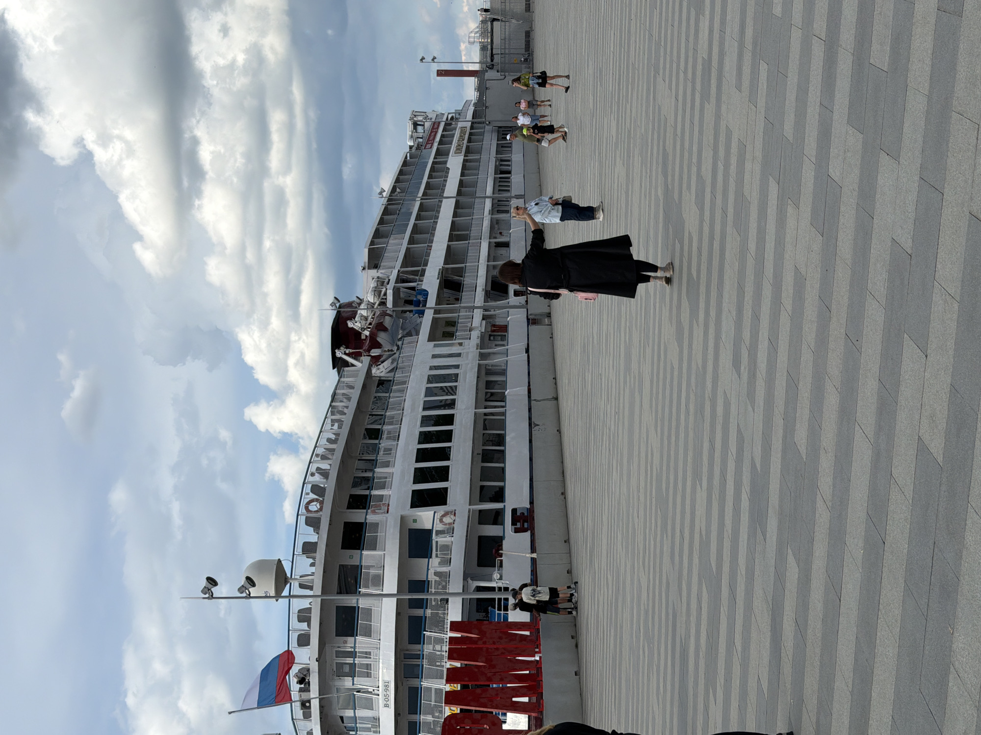 A wide shot shows a large white passenger ship docked at a waterfront promenade in Perm, Russia. The ship, identified by the writing "PAM" in large red letters on its side, appears to be a river cruise vessel with multiple decks, windows, and balconies. A Russian flag flies from the top deck.
The promenade in the foreground is paved with grey and lighter grey rectangular tiles laid in a patterned fashion. Several people are walking on the promenade. In the center of the frame, a woman in a black coat and leggings, with a pink bag slung over her shoulder, is walking away from the camera, gesturing with her right hand. To her left, two young males are standing near the red letters, one on a scooter. Further in the distance, a group of people, including a woman in a light blue top and jeans, a man in a green shirt and shorts with a child in a yellow hat, and a woman in a floral dress, are also walking on the promenade.
The sky is cloudy with patches of blue. The overall scene depicts a public area by the water on a cloudy day.