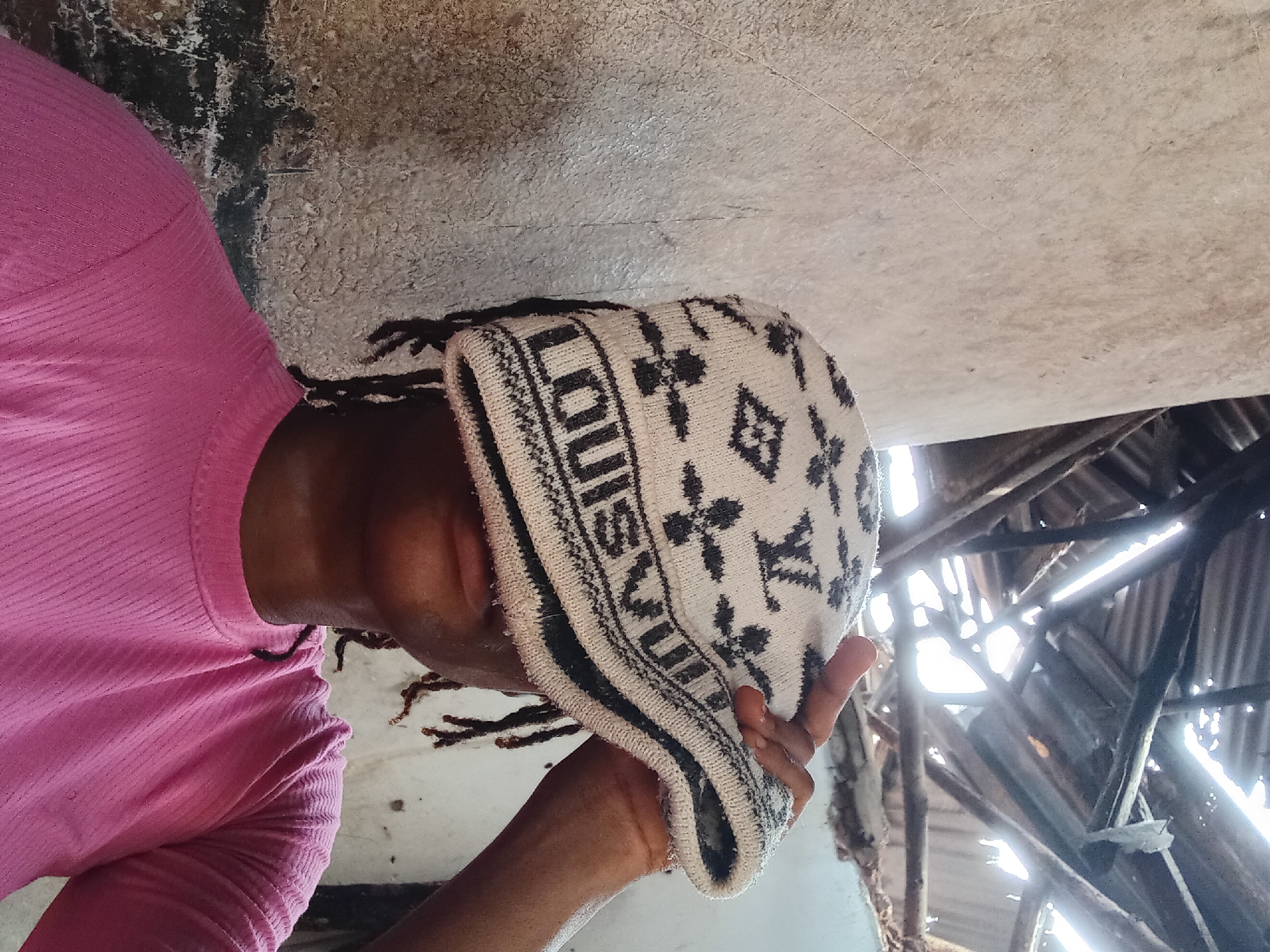 A person, likely of Black ethnicity, is depicted in a close-up, rotated image, wearing a pink long-sleeved shirt and a patterned knitted beanie. The individual's face is mostly obscured as the beanie is pulled low, covering their eyes and forehead, and they are looking slightly upwards or to the side. Visible features include parts of their dark-skinned neck and chin, and dark dreadlocks or braids extending from beneath the beanie.
The beanie is cream or off-white with black patterns, prominently featuring the word "LOUIS" on its brim and repeating stylized "V" or flower-like motifs, highly suggestive of the Louis Vuitton monogram. The person's left hand is visible, gently holding the brim of the beanie near their chin.
The background is rustic and appears to be an interior or covered outdoor space. To the left and above the person, light-colored walls or ceiling sections show signs of age, dirt, or mold. On the right side of the frame, behind and above the person's head, there's a clear view of a damaged or very basic roof structure. This structure consists of wooden beams and corrugated metal sheets, possibly rusted or charred, through which bright daylight streams in, creating strong highlights and shadows. The overall impression of the setting is one of an old, neglected, or perhaps unfinished building. The bright light indicates it is daytime.