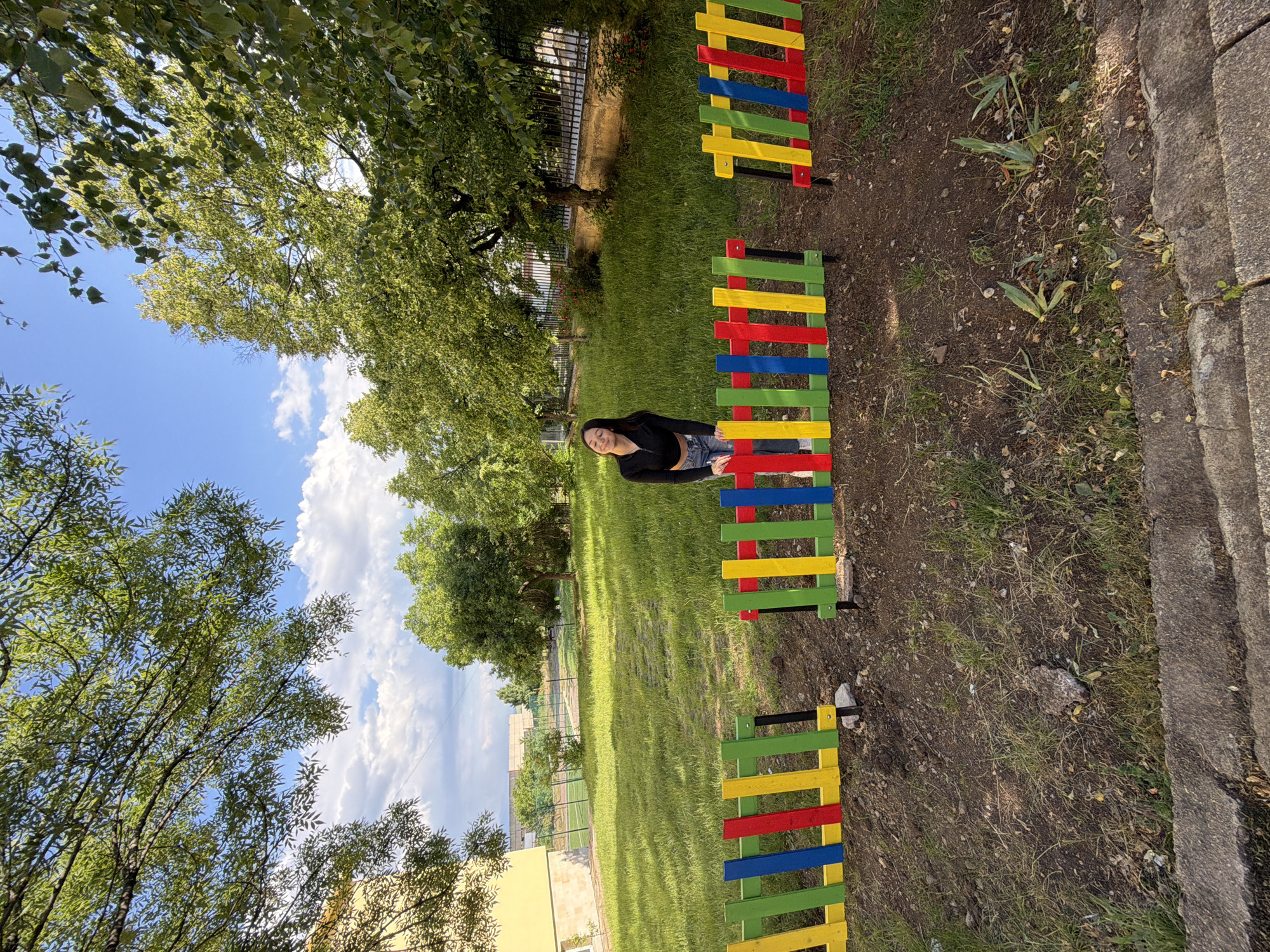 This outdoor image captures a young woman seated on a colorful, low barrier in a park-like setting. The barrier consists of horizontal slats painted in alternating bright red, blue, yellow, and green, suggesting a decorative or play-area element. The woman, wearing a dark top and blue jeans, faces the camera with a relaxed posture and a soft smile.
The scene is dominated by lush greenery. A wide expanse of mown grass extends behind the woman, leading to more trees and a metal fence in the mid-ground. Beyond the fence, what appears to be a sports field or track and a light-colored building are visible. A dirt path runs alongside the colorful barriers, eventually meeting a concrete curb or sidewalk on the right side of the frame. Tall, leafy trees with vibrant green foliage frame the upper and left portions of the image. The sky is bright blue with scattered white clouds, indicating clear and sunny weather, likely late morning or afternoon given the natural light and shadows. The overall impression is one of leisure and pleasant outdoor activity on a beautiful day. No text is visible in the image.
