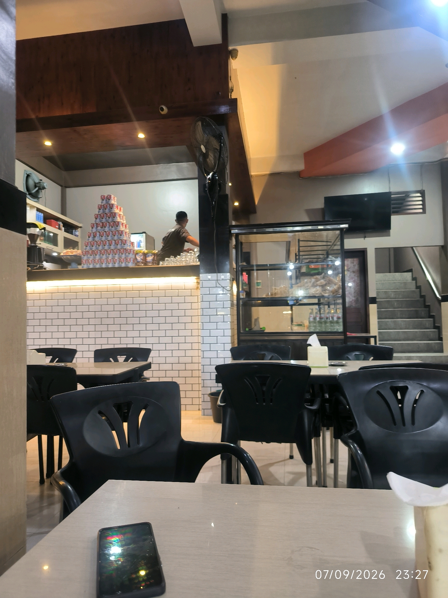 The image captures the interior of a restaurant, likely located in Keudebayu, Indonesia, with a date and time stamp of 07/09/2026 23:27. In the foreground, a smartphone lies on a light-colored table. Behind the table, several black plastic chairs with distinctive V-shaped cutouts are visible, arranged around tables. The background reveals a counter area with a stack of canned goods, possibly condensed milk, arranged in a pyramid. A person, seen from the back, is working behind the counter. White subway tiles line the wall behind the counter. Further in the background, a glass display case showcases food items, and a staircase is visible on the right. A black oscillating fan is mounted on the wall near the ceiling. The overall ambiance suggests a casual dining establishment.