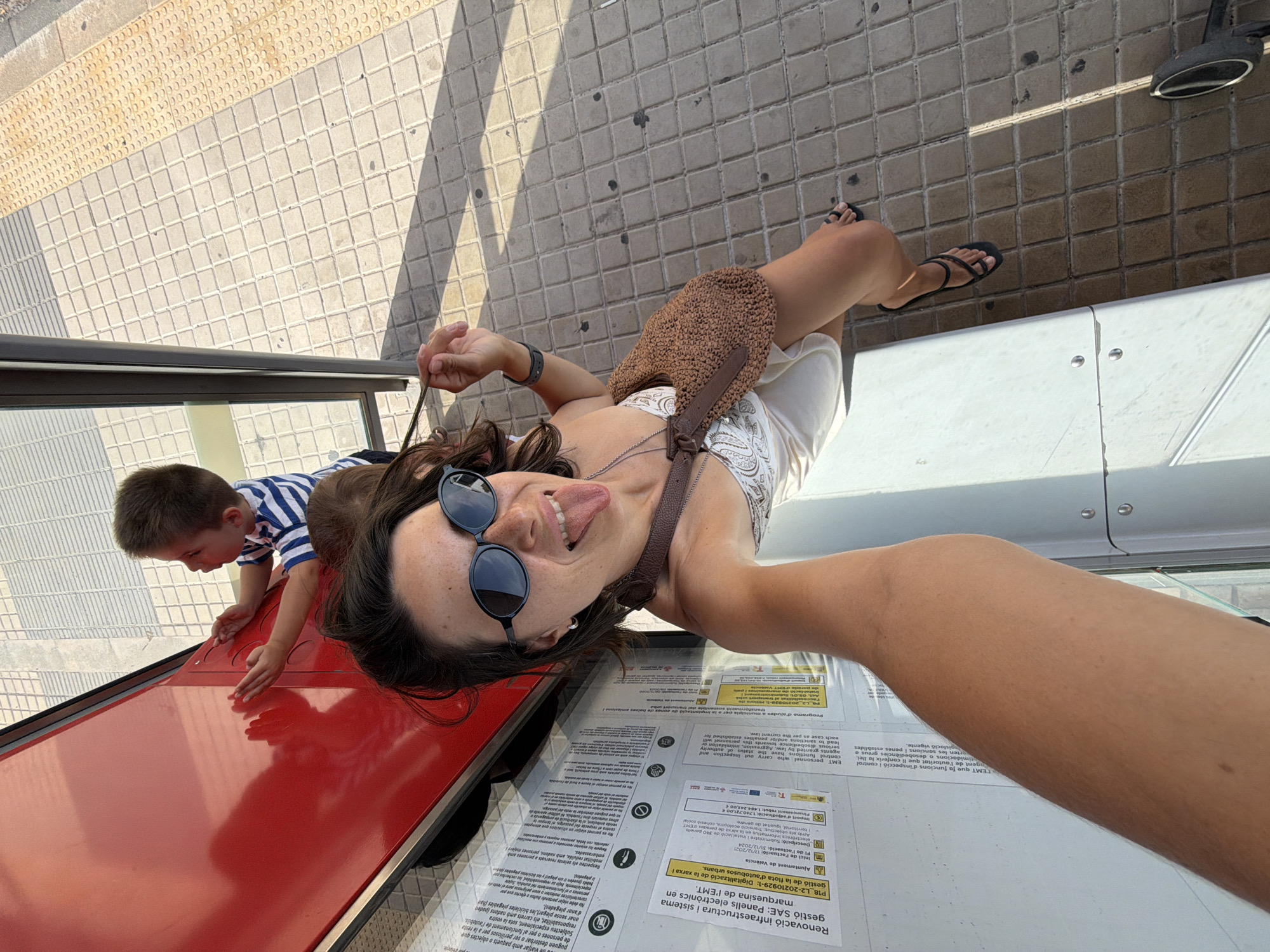 A high-angle, full-body selfie captures a woman with dark hair and sunglasses playfully sticking her tongue out at the camera. She wears a white top with a patterned detail and a brown woven crossbody bag. Her right hand is held near her face, while her left arm is outstretched to capture the selfie. Beside her, a young boy in a striped blue and white shirt is seen from the back, looking at a bright red surface with textured indentations. The scene appears to be outdoors, possibly at a public transit stop, with a tiled ground and a yellow tactile paving strip visible. Information posters are affixed to a vertical surface on the left side of the frame. The lighting suggests a sunny day, casting shadows on the ground.