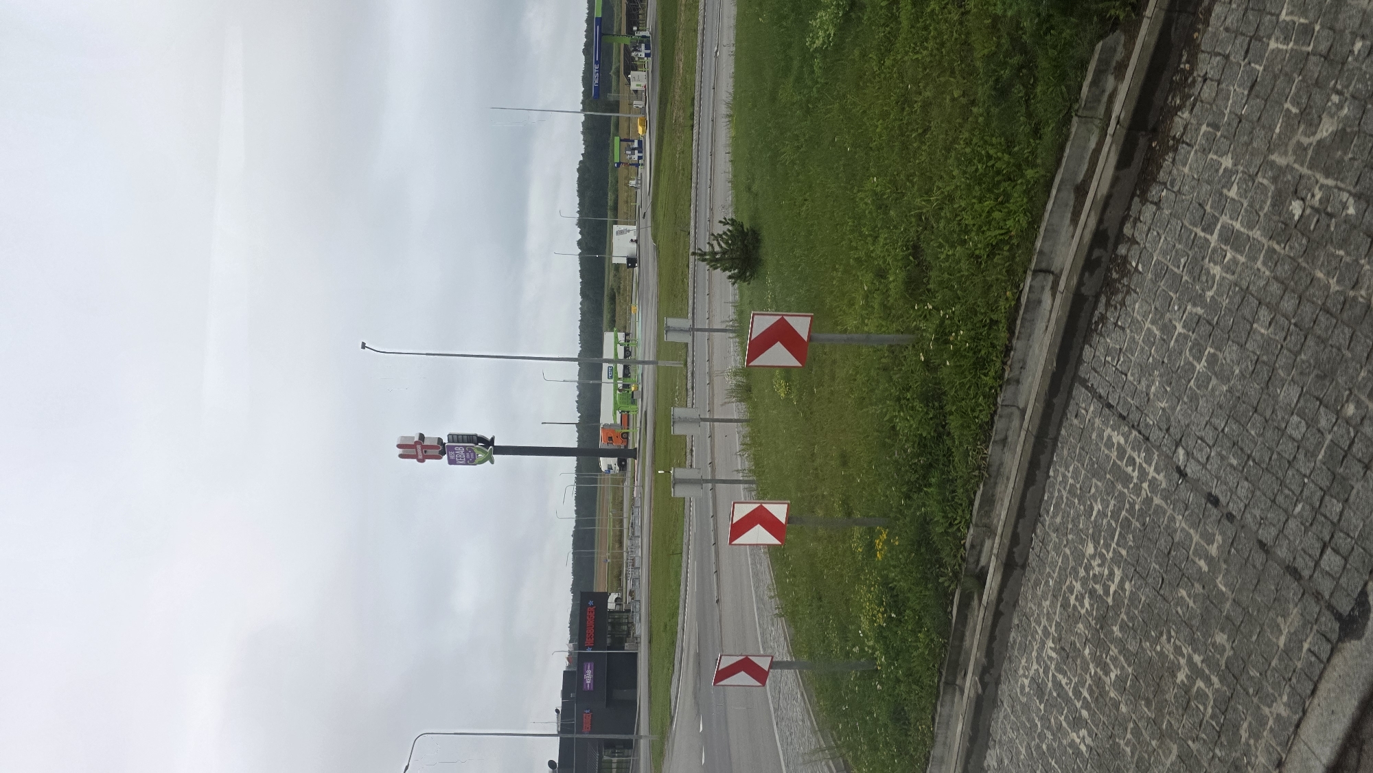 The image shows a cloudy day scene at what appears to be a roadside service area or gas station. In the foreground, a cobblestone path curves to the left, bordered by a concrete curb and a grassy area with several small white and red directional signs indicating a curve to the right. In the mid-ground, a building with a sign reading "HESBURGER" is visible, suggesting a fast-food establishment. Across the road, a green and blue gas station with "NESTE" branding is present, with fuel pumps and a yellow vehicle. A tall sign with a red "H" logo and text "KEBAB" is also prominent. Further in the background, there are trees and fields. The overall atmosphere is overcast, with no clear sky visible. The location is identified as Pamolainiai, Lithuania.