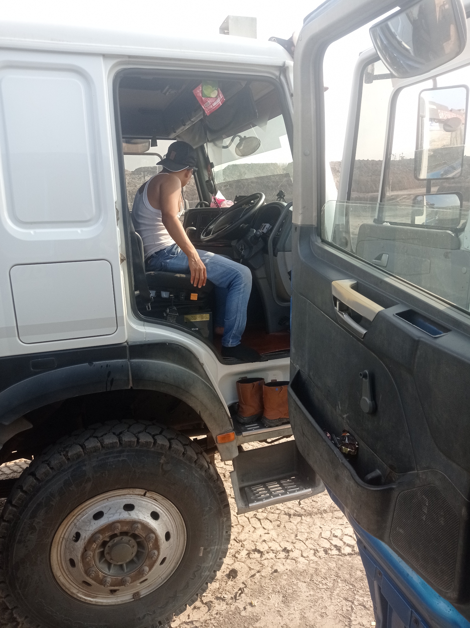 A man wearing a white tank top, blue jeans, and a black baseball cap sits in the driver's seat of a white semi-truck. The driver's side door is open, revealing the interior of the cab. The man's right hand rests on the steering wheel. Two brown work boots are visible on the floorboard inside the truck. Outside the truck, a large tire is prominent in the foreground. The ground is covered with dry, cracked earth. The scene appears to be in an open area, possibly a construction site or a rural location, with dirt piles and some distant structures visible in the background. The lighting suggests it is daytime, with bright sunlight casting shadows. The image was taken at an angle, looking into the truck cab from the right side.