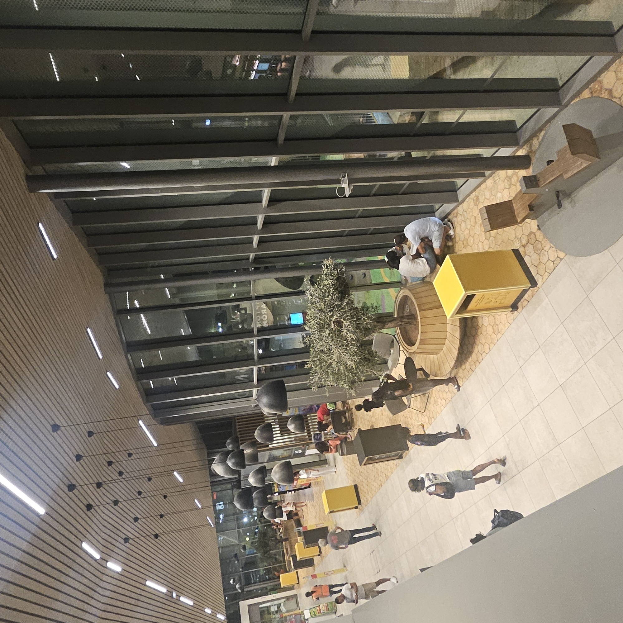The image shows an interior view of a modern building, likely a public space such as a shopping center or an airport terminal, in Lançon-Provence, France. The architecture features a sloped wooden-slatted ceiling with linear lighting and large glass windows overlooking an outdoor area with some greenery and reflections of lights. Inside, the floor is tiled with large, light-colored square tiles, interspersed with areas of hexagonal stone tiles. Several black, bell-shaped pendant lights hang from the ceiling, creating a decorative element.
On the left side of the image, a walkway is visible, with a few people present. One person, a woman wearing a white top and denim shorts, walks away from the camera carrying a dark bag. Another person, a man in a white t-shirt and shorts, stands looking at his phone. Further down the walkway, a person in an orange vest is seen walking. Yellow bins are placed along the walkway.
In the central area, an artificial tree in a large wooden planter serves as a focal point. Around the tree, there are seating areas with wooden benches. A group of people, two women and a man, are seated on the floor near the tree, interacting. Another yellow bin is positioned nearby, along with a wooden seesaw-like playground structure. The glass facade of the building reveals a glimpse of the outdoor environment, showing some lighting and possibly more trees or landscaping. The overall impression is one of a bright, contemporary, and somewhat spacious public area.