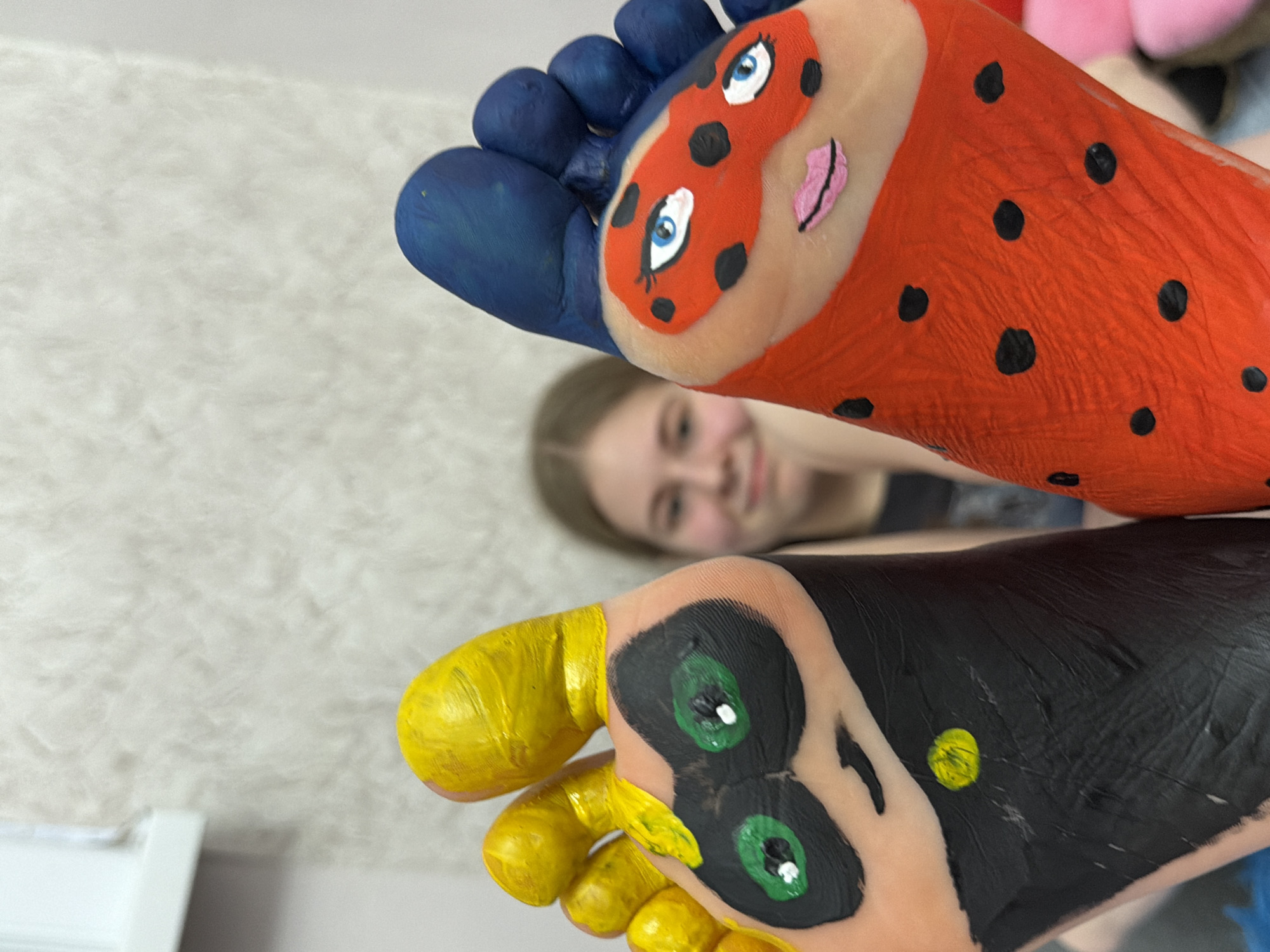 A close-up indoor shot features two feet painted to resemble characters, with a blurred person in the background. The top foot is painted red with black polka dots, blue toes, and a cartoon face with blue eyes and pink lips. The bottom foot is painted black with yellow toes, green eyes, and a small yellow spot. The person in the background is slightly out of focus and appears to be smiling. The location is potentially in Perm, Russia, but the photo itself does not contain any evidence to confirm this location. The environment includes a soft, off-white surface, possibly a rug, and part of a white object in the lower left corner.