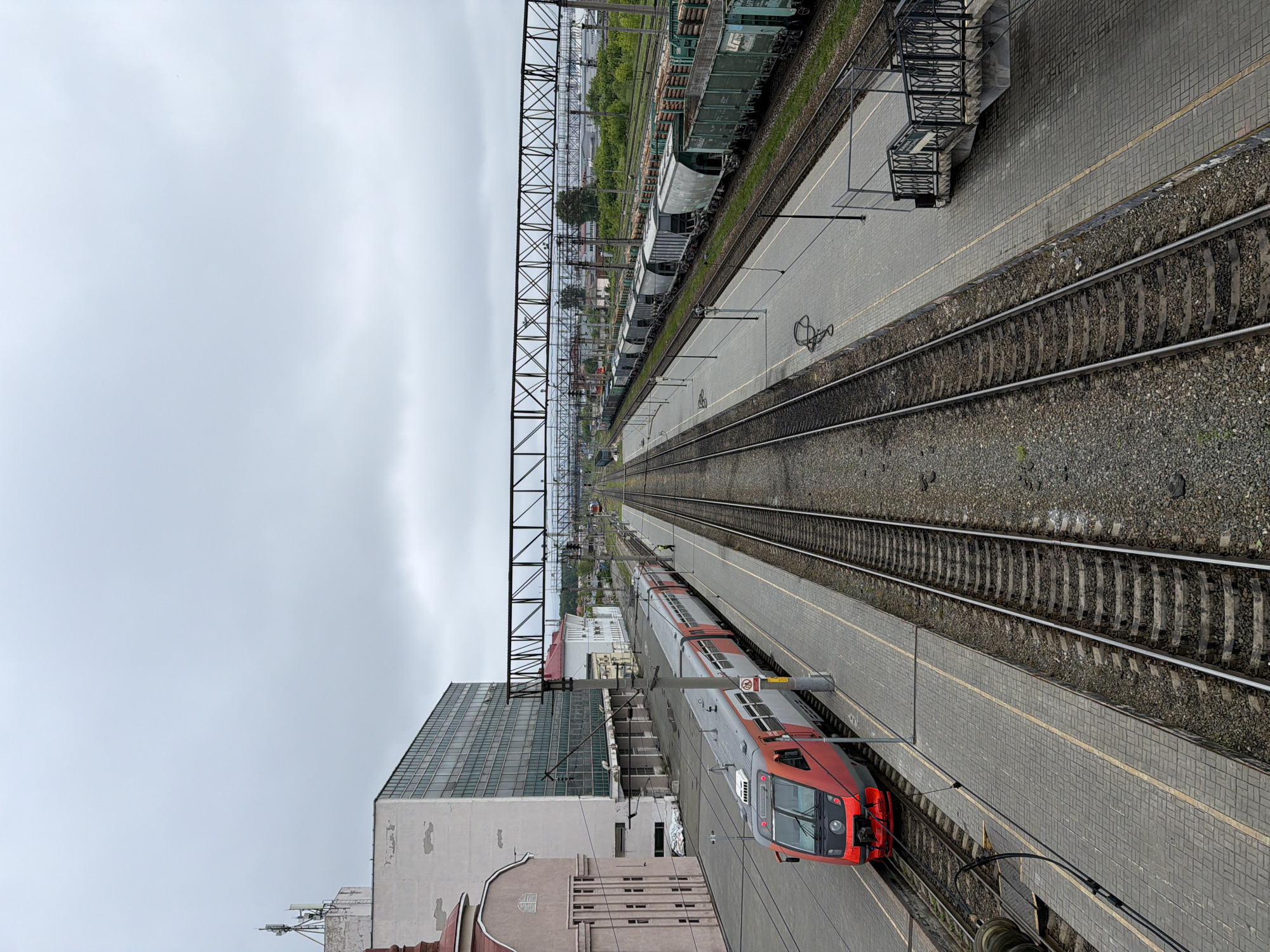 This image provides an elevated, overhead view of a train station and railway lines in Izhevsk, Russia, on an overcast day. The perspective looks along the length of multiple parallel tracks extending into the distance.
In the foreground and midground, a modern red and silver passenger train is parked at a paved platform on the left side of the frame. The platform features a yellow safety line along its edge. To the immediate left of the tracks and platform, a large building with a contemporary glass facade stands tall, flanked by parts of older buildings. Numerous overhead electrical lines and substantial metal support structures (gantries) crisscross above the tracks.
Further to the right, several more train tracks are visible, laid on a bed of gravel ballast, along with a second platform. In the background, on tracks further afield, various freight wagons are parked; notably, several green wagons are loaded with timber, alongside other grey cargo cars. Beyond the extensive railway yard, a scattering of trees and urban structures completes the distant horizon.
The scene appears quiet, with no individuals clearly visible on the platforms or around the trains. The sky is uniformly cloudy, suggesting a dull daytime. Visible text on a yellow warning sign attached to an overhead pole near the passenger train reads "ОСТОРОЖНО ВЫСОКОЕ НАПРЯЖЕНИЕ" (CAUTION HIGH VOLTAGE) and "НЕ ЗАХОДИТЬ ЗА ЖЕЛТУЮ ЛИНИЮ" (DO NOT GO BEHIND THE YELLOW LINE). Additionally, "С О Р Т О В О Й" is partially legible on one of the green freight wagons.