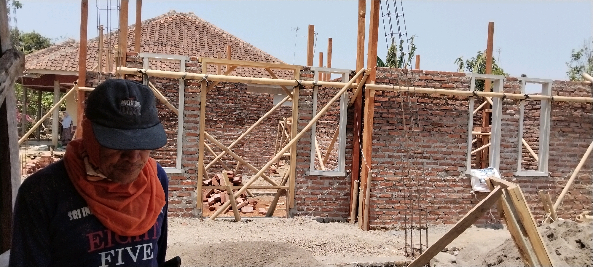 The image captures a construction site in Pamulihan, Indonesia, where a house is being built. In the foreground, a man wearing a dark blue t-shirt with "EIGHT FIVE" printed on it and an orange scarf is looking down. He is also wearing a dark baseball cap with the logo "MICHELO DRY". Behind him, the brick walls of a house under construction are visible, with wooden scaffolding and framing in place. Two window frames are installed in the brick wall. In the background, the tiled roof of an existing structure and some trees can be seen, along with a clear sky. The ground is covered with gravel and some piles of bricks. The overall scene depicts an active construction project with one individual prominently featured in the foreground.