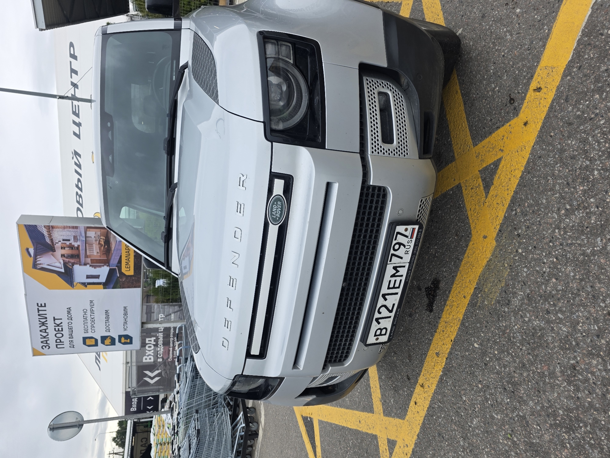 The image shows a silver Land Rover Defender parked on asphalt marked with yellow lines. The vehicle's license plate reads "B121EM797 RUS". Behind the vehicle, a large white banner with Russian text is visible, advertising a service called "LEMANAPE" related to "PROJECTS FOR YOUR HOME" including free design, delivery, and installation. To the left, shopping carts are stacked, and another banner reads "ВХОД В САДОВЫЙ ЦЕНТР" (Entrance to the Garden Center). The scene appears to be an outdoor commercial area. The specific location is identified as Sholokhovo, Russia. There are no people visible in the image.
