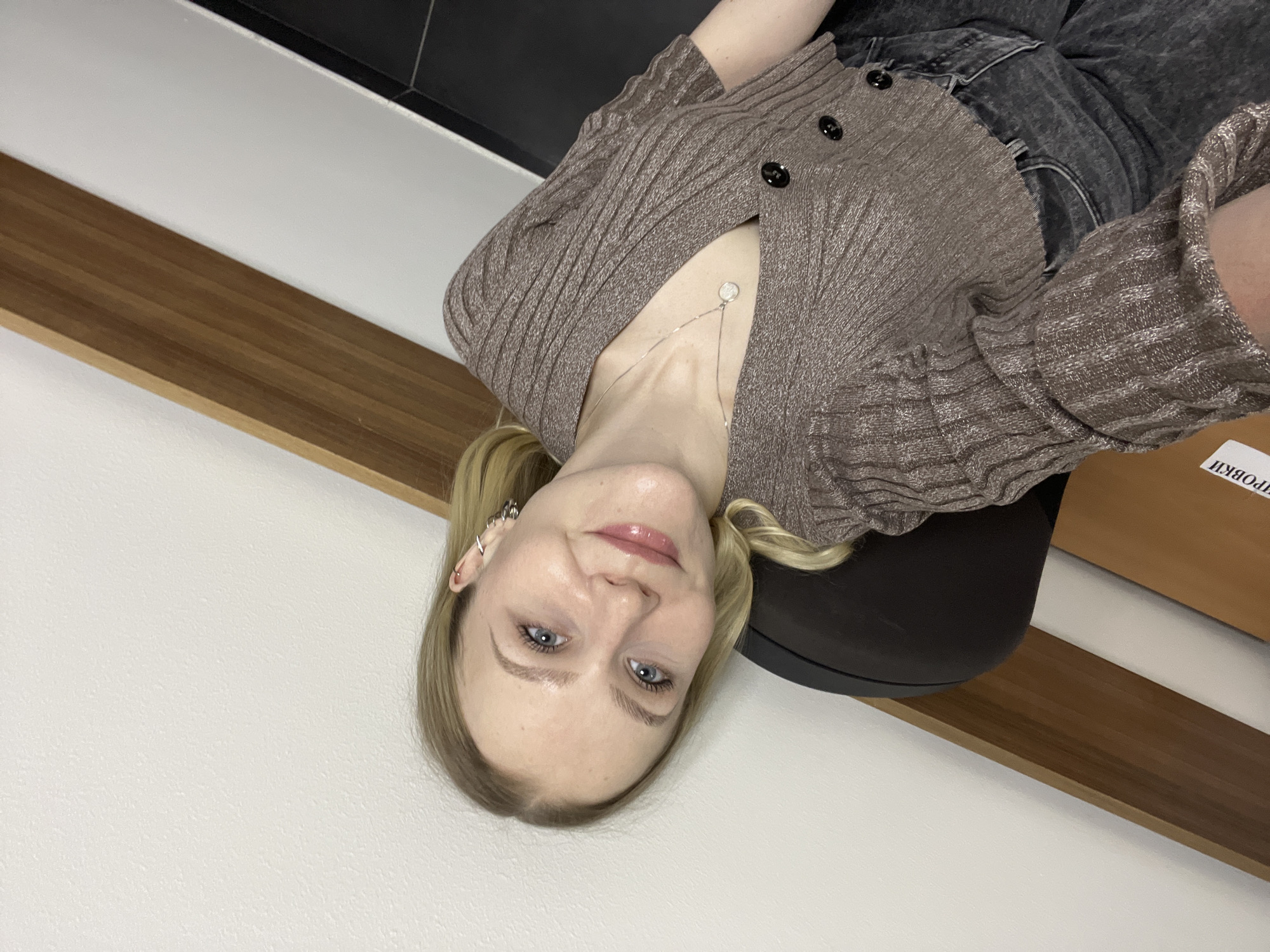 A young woman with blonde hair and blue eyes poses for a photograph. She is wearing a brown cardigan and a silver necklace. She is seated on a black chair in what appears to be an office or interior setting. In the background, there is a wooden shelf, a white wall, and a dark surface above. The inscription "ПРОВКИ" is visible on a wooden surface. There is no verifiable information to confirm the location is Khimki, Russia.